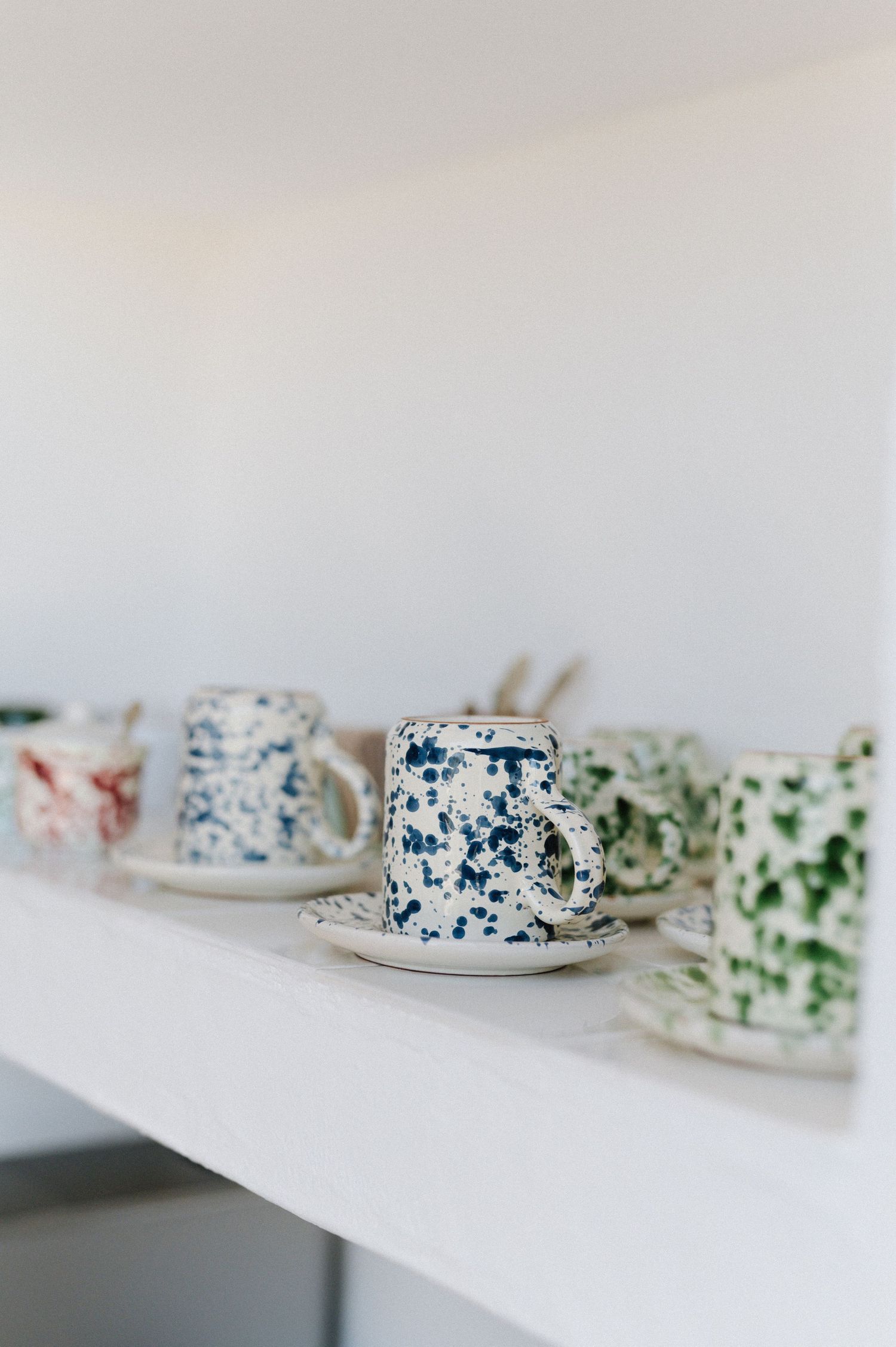 Speckled ceramic coffee cups and saucers with colorful splatter patterns displayed on a white shelf.