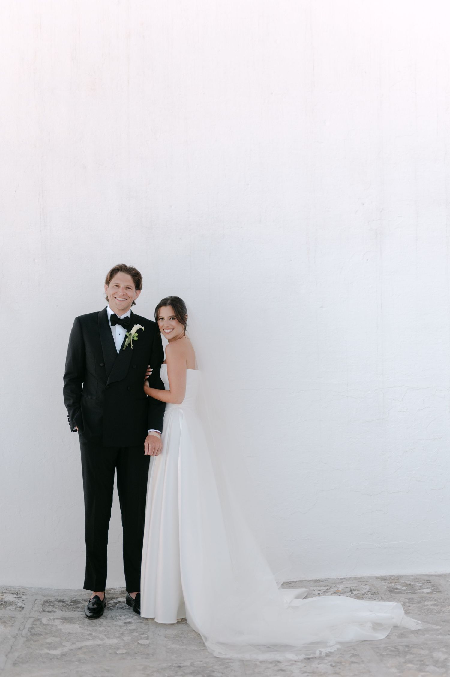 Bride and groom posing together against a white wall, groom in black tuxedo, bride in elegant white gown.