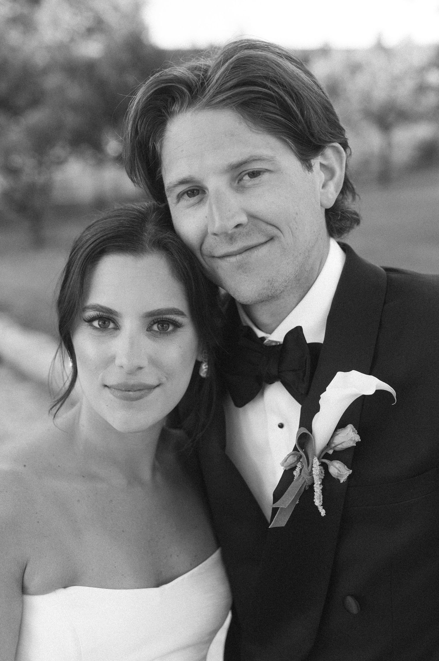 Black and white wedding portrait of smiling bride and groom in tuxedo with boutonniere posing together outdoors.