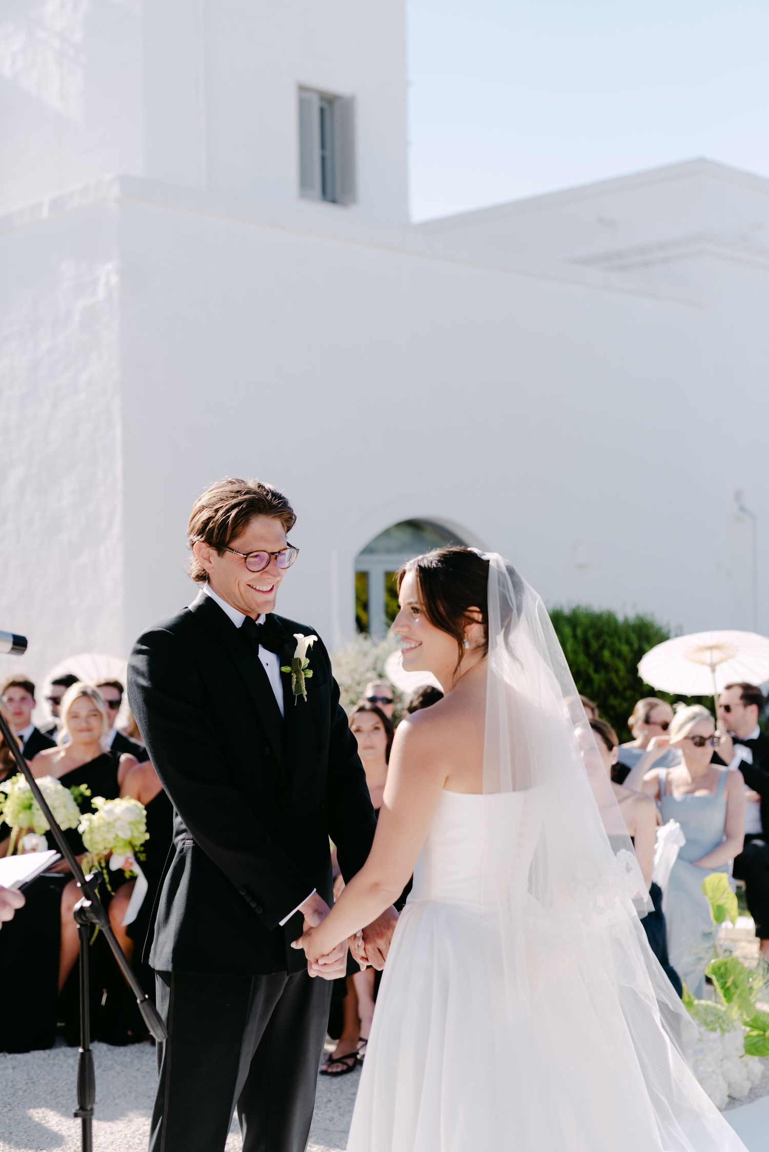 Bride and groom holding hands at outdoor wedding ceremony beside white building on sunny day.