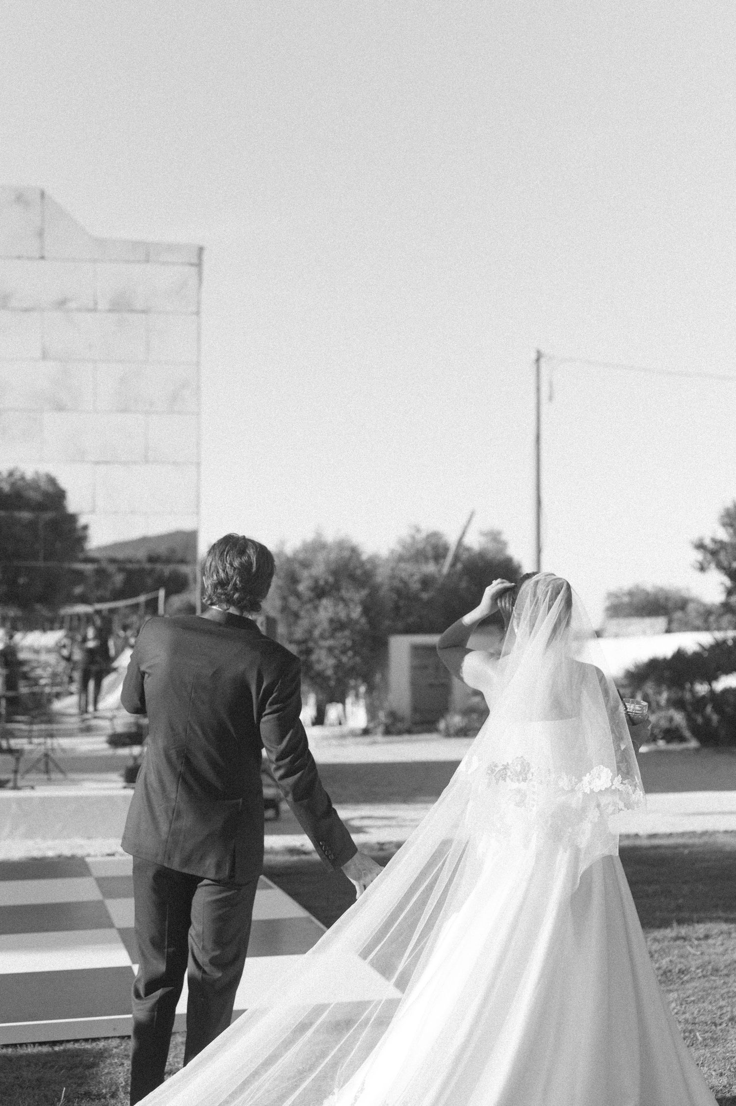 Black and white photo of a bride and groom walking hand in hand, her veil flowing in the breeze at an outdoor wedding.