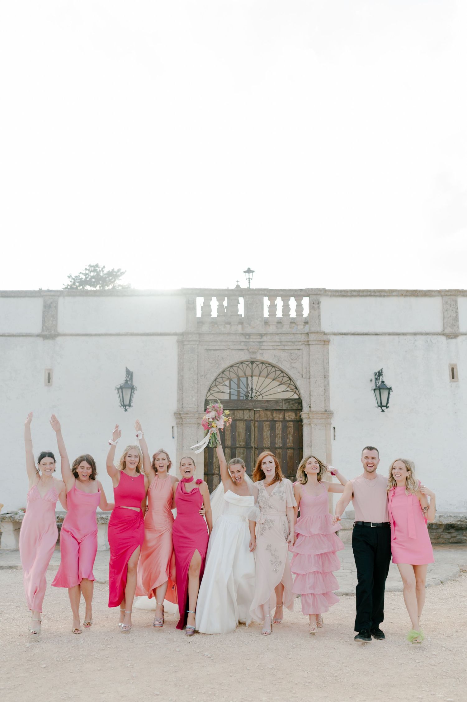 Wedding party in pink and white dresses poses together in front of elegant white stone archway entrance.