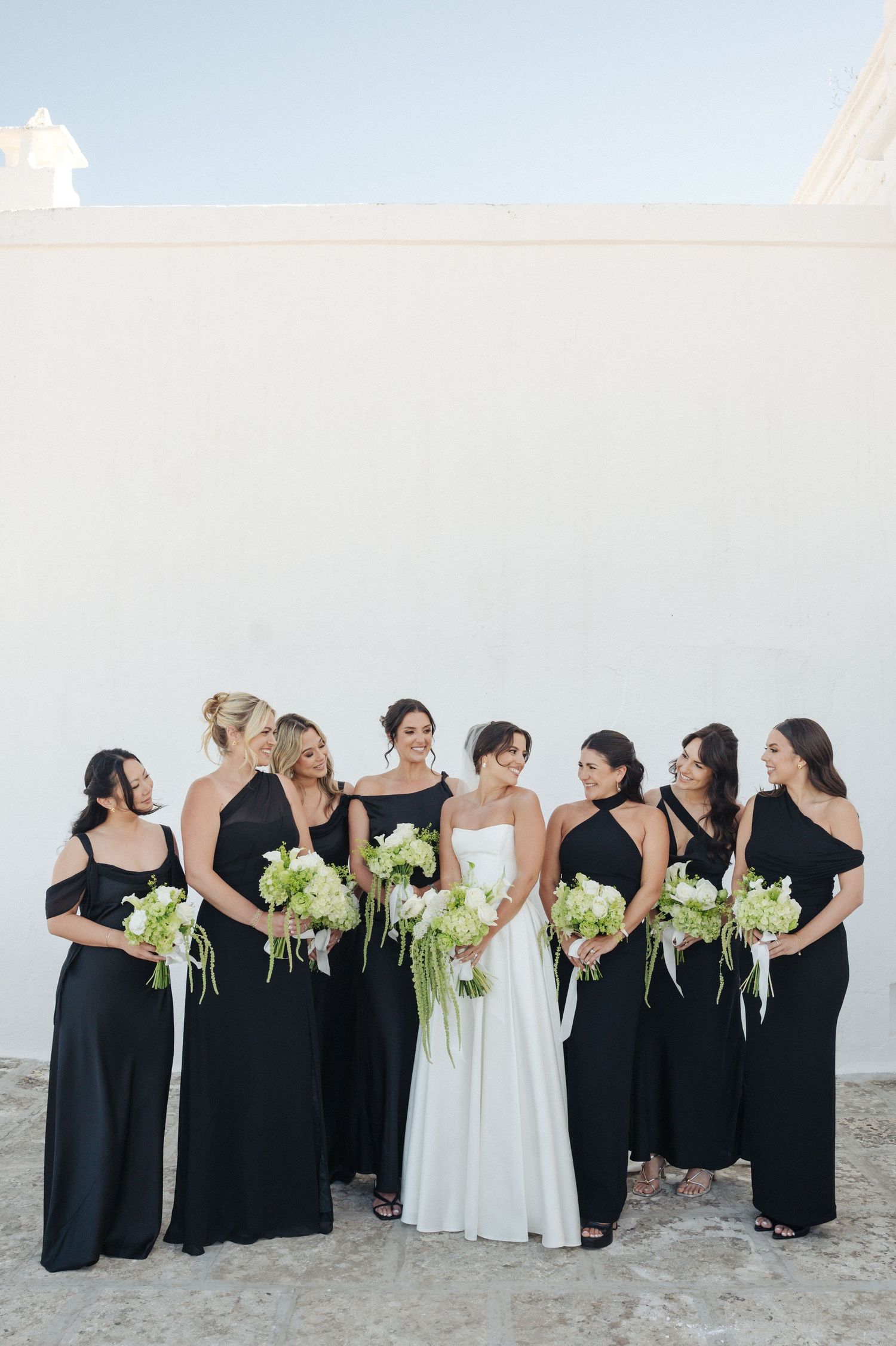 Bride in white gown with six bridesmaids in black dresses holding white floral bouquets against a white wall.