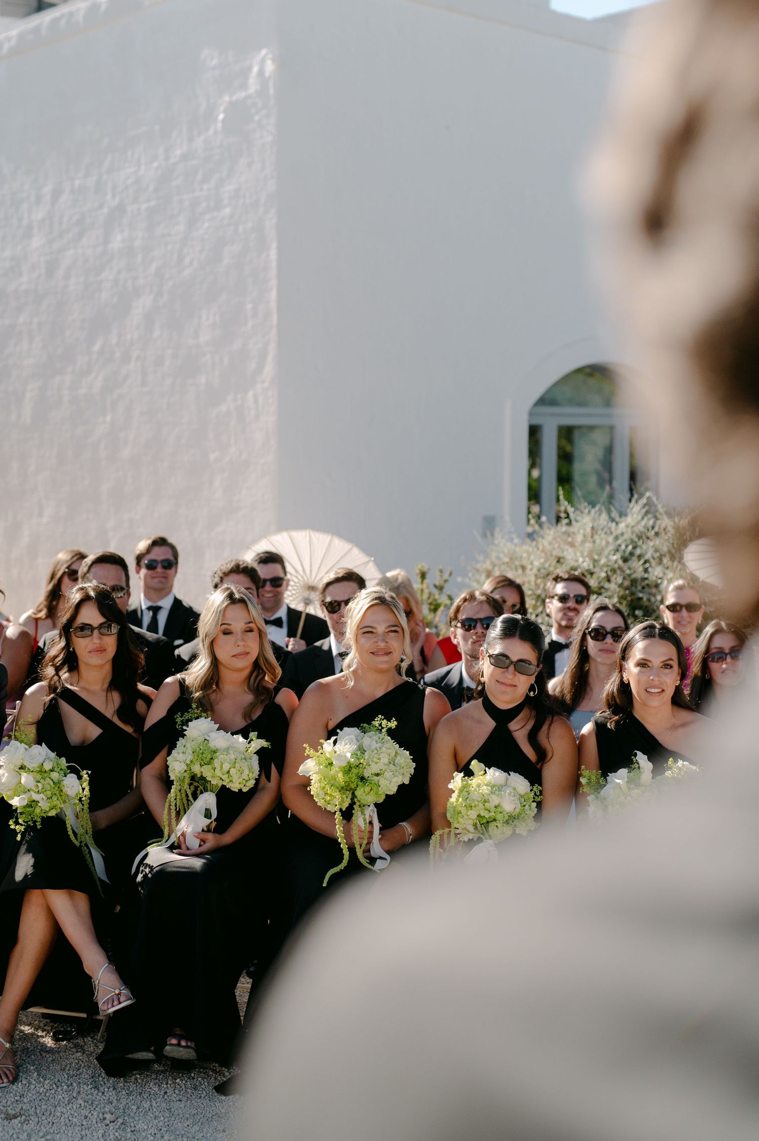 Bridesmaids in black dresses holding white floral bouquets seated at an outdoor wedding ceremony.