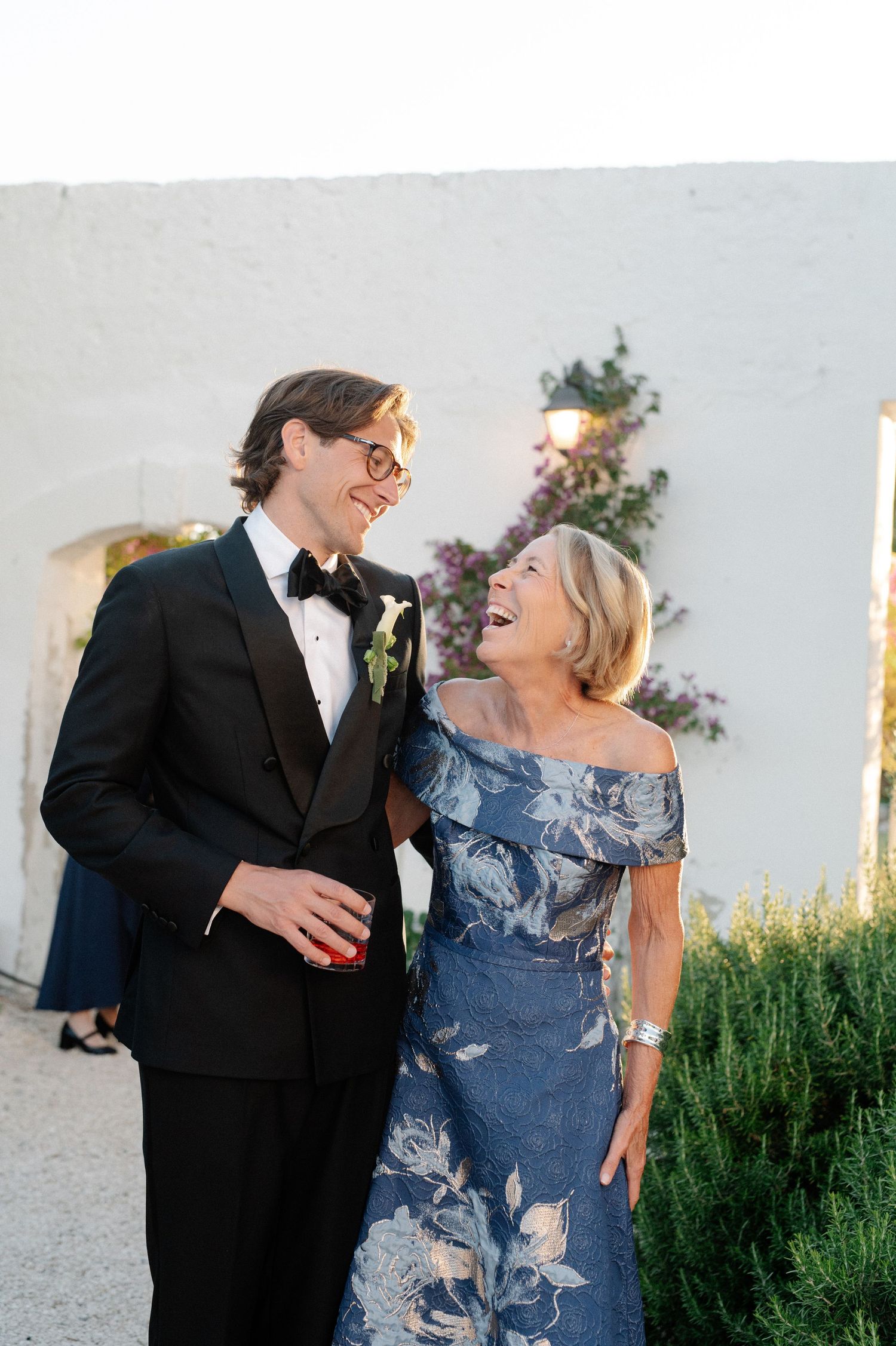 Groom in black tuxedo laughing with older woman in blue floral off-shoulder dress at outdoor wedding venue.