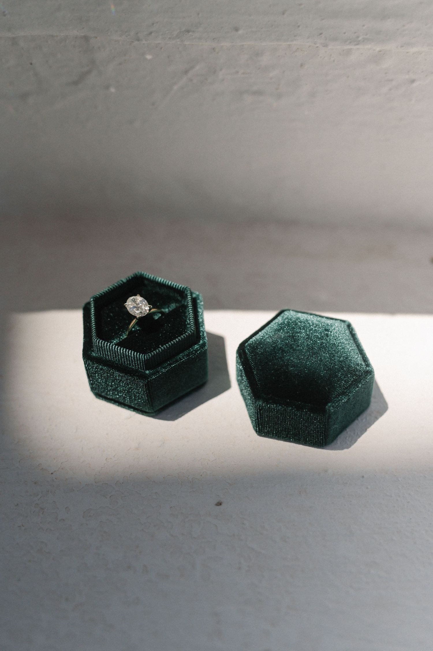 Dark green hexagonal velvet ring box open displaying a diamond solitaire engagement ring on white surface.