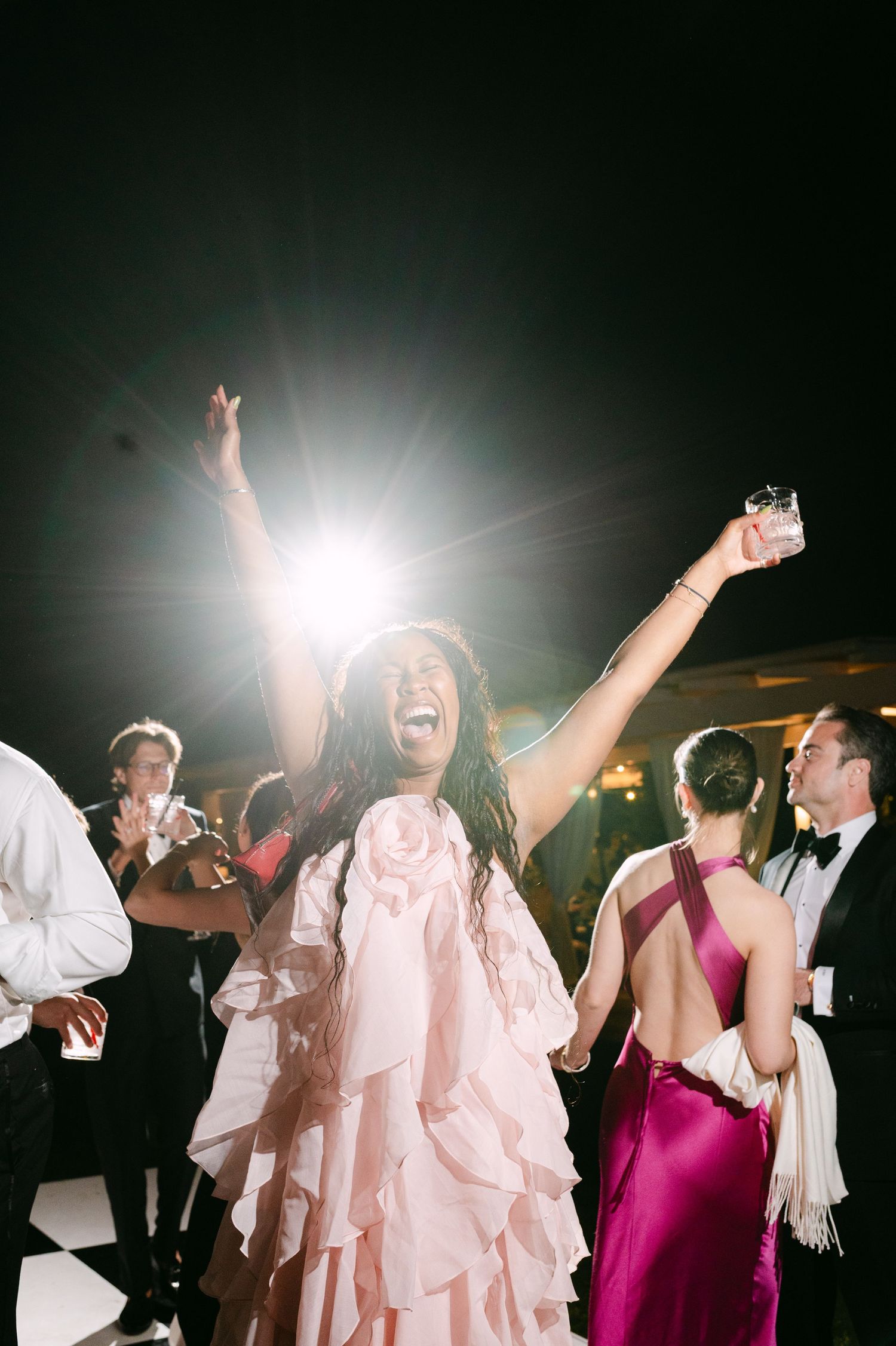 A joyful woman in a pink ruffled gown dances with raised arms at an elegant evening wedding reception.