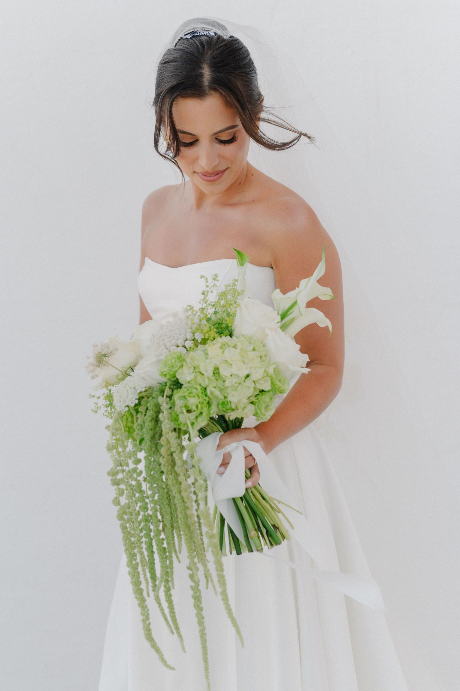 Bride in strapless white gown holding lush green and white floral bouquet with cascading amaranthus, posing against white wall.