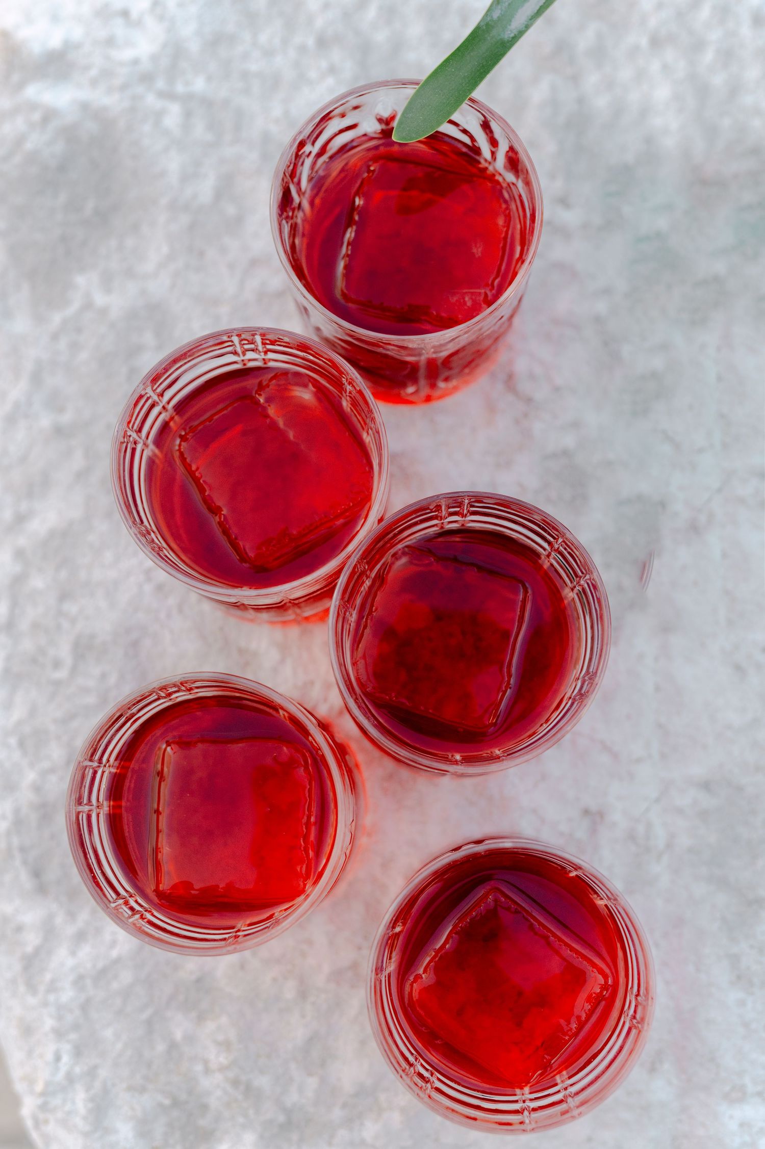 Five glasses of red iced drinks arranged on a marble surface, one with a green straw.