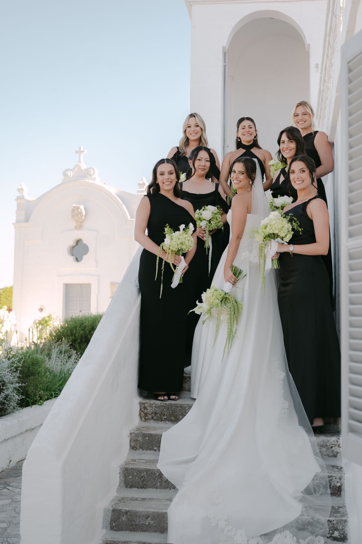 Bride in white gown with bridesmaids in black dresses holding white bouquets on church steps in Santorini.