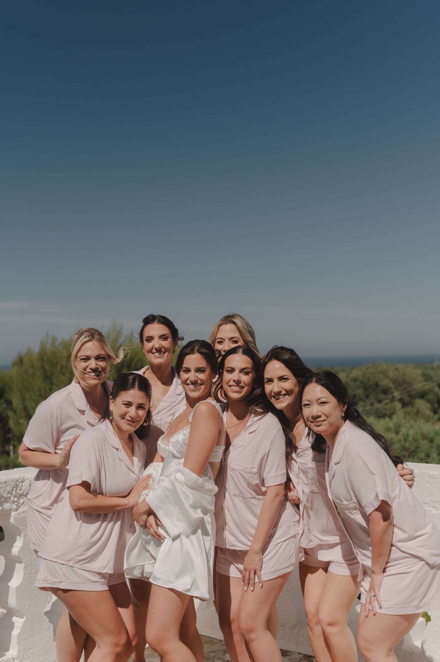 Bride and bridesmaids in matching pink pajamas posing together outdoors on a sunny day.