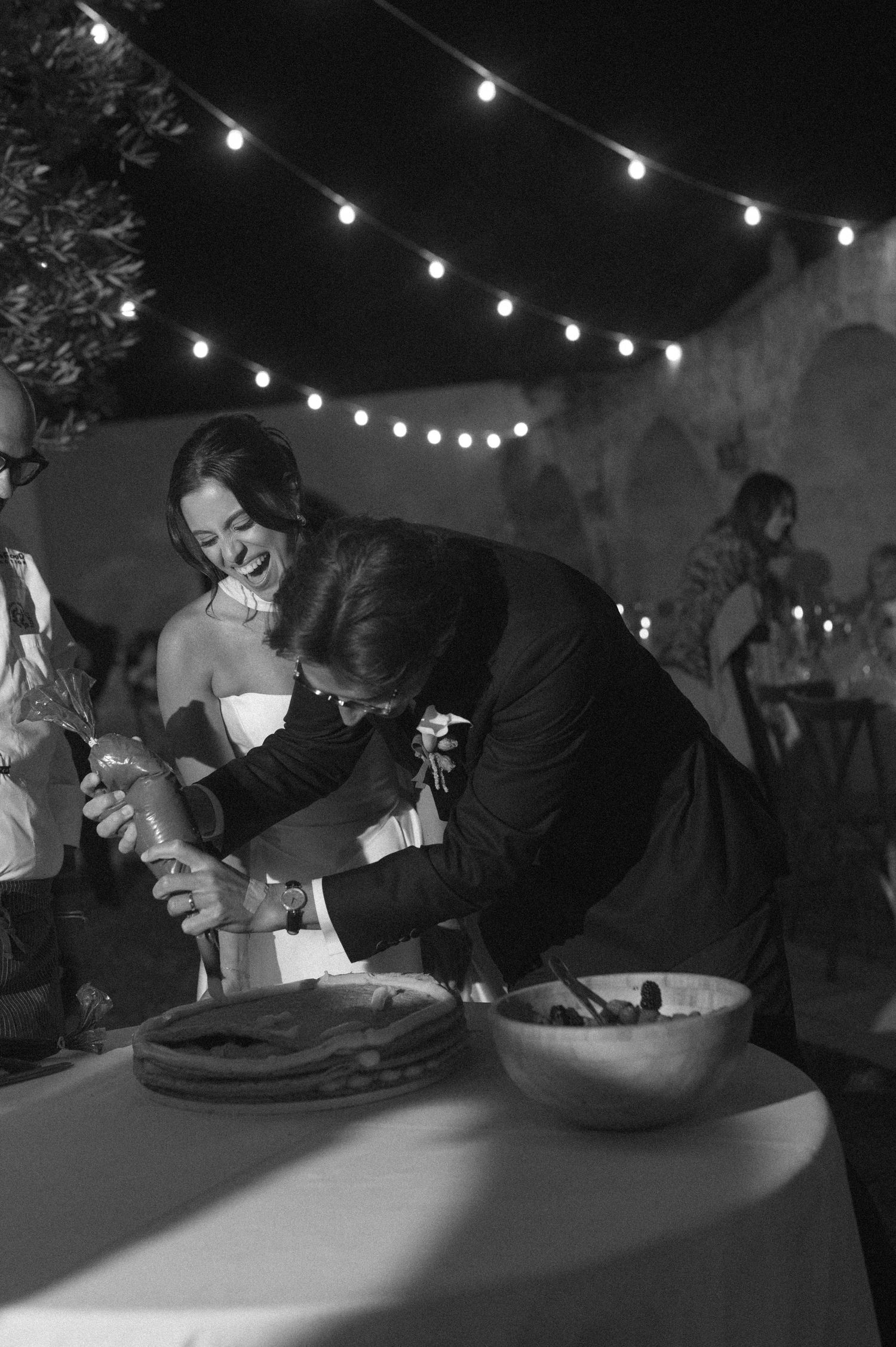 Black and white photo of a bride and groom laughing while cutting their wedding cake under string lights at night.