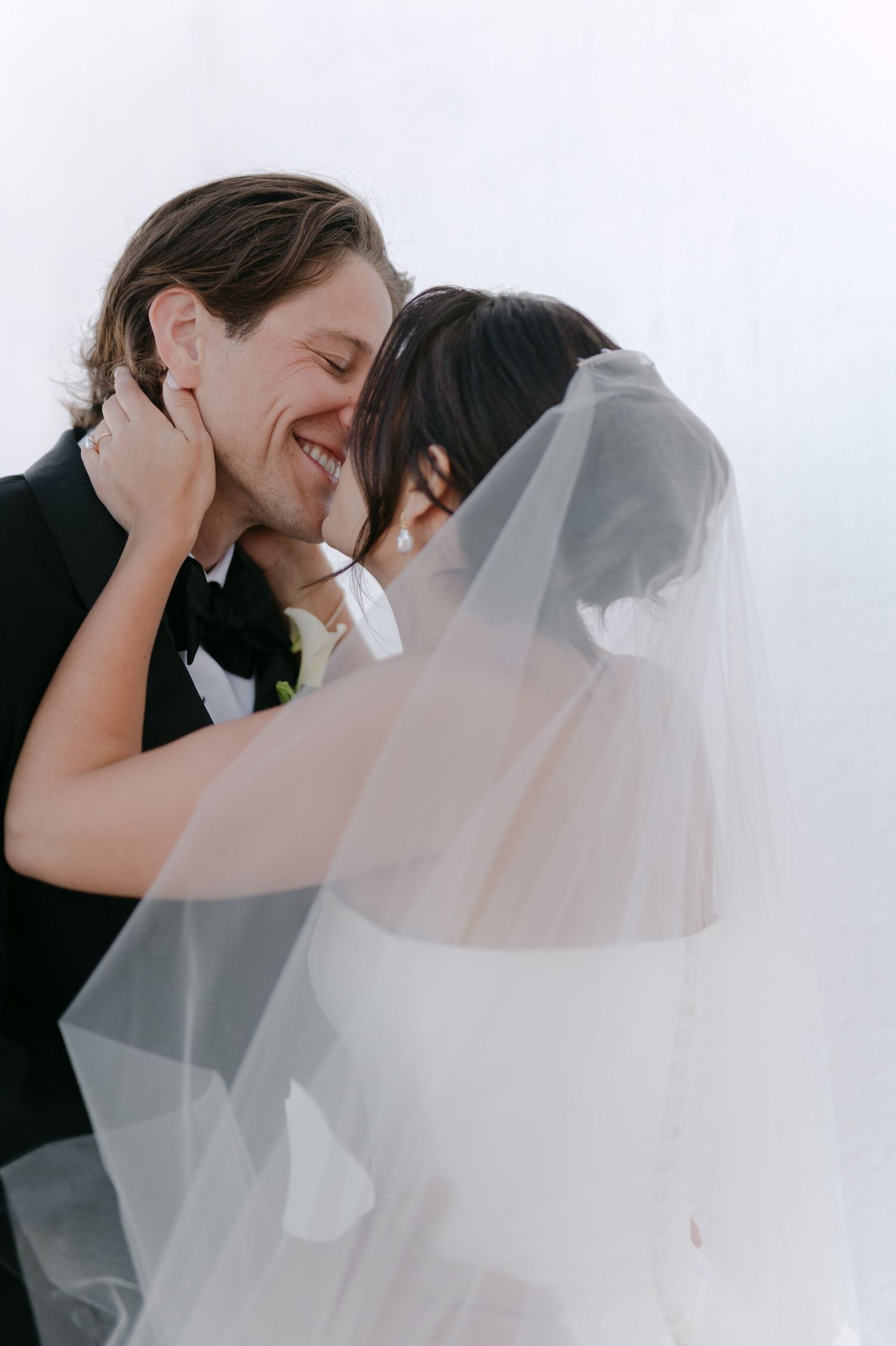 A smiling groom in a black tuxedo shares a romantic moment with his veiled bride on their wedding day.