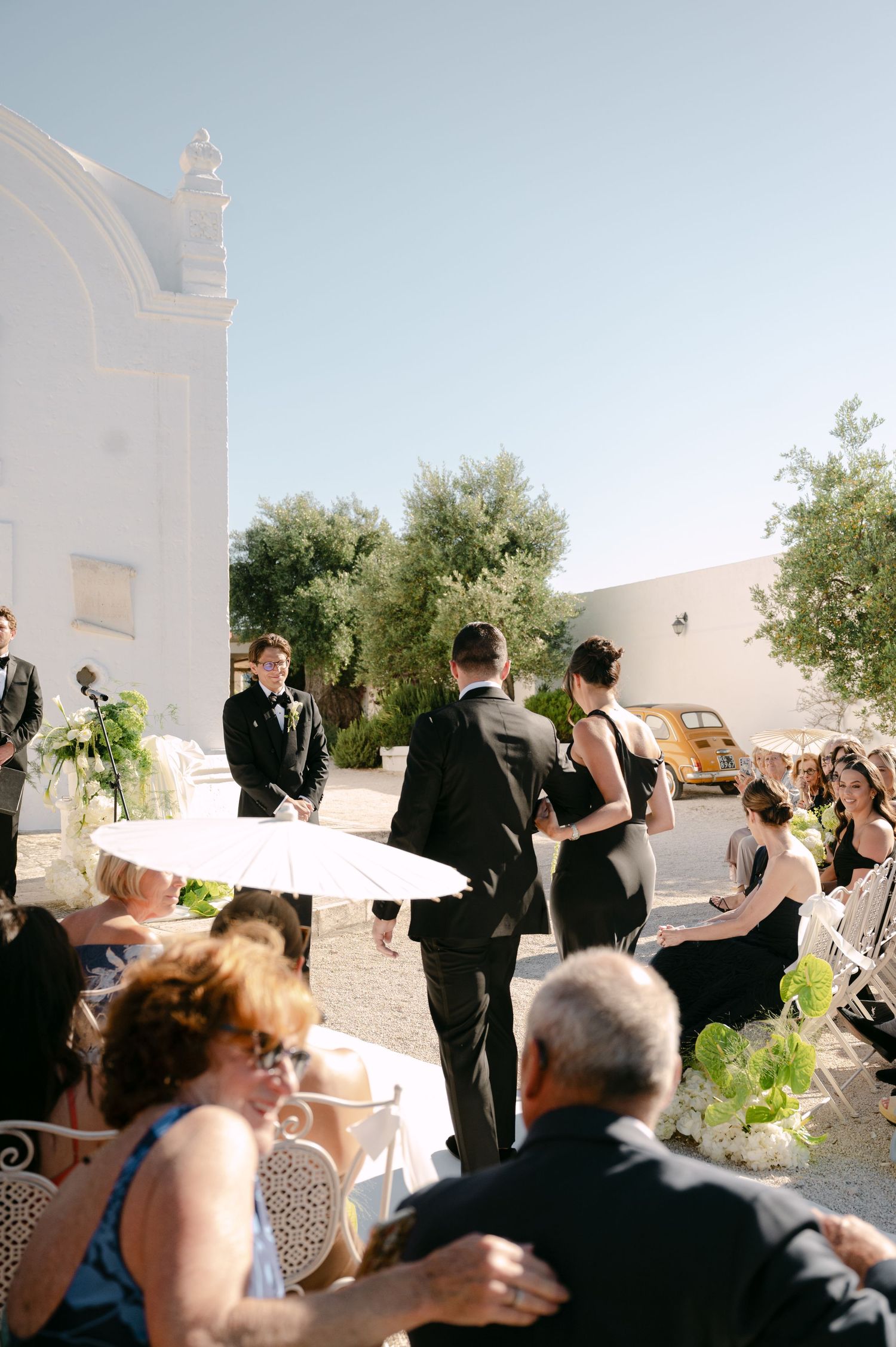 Outdoor wedding ceremony with guests seated in white chairs, bridal party in black attire near a white church, sunny day.