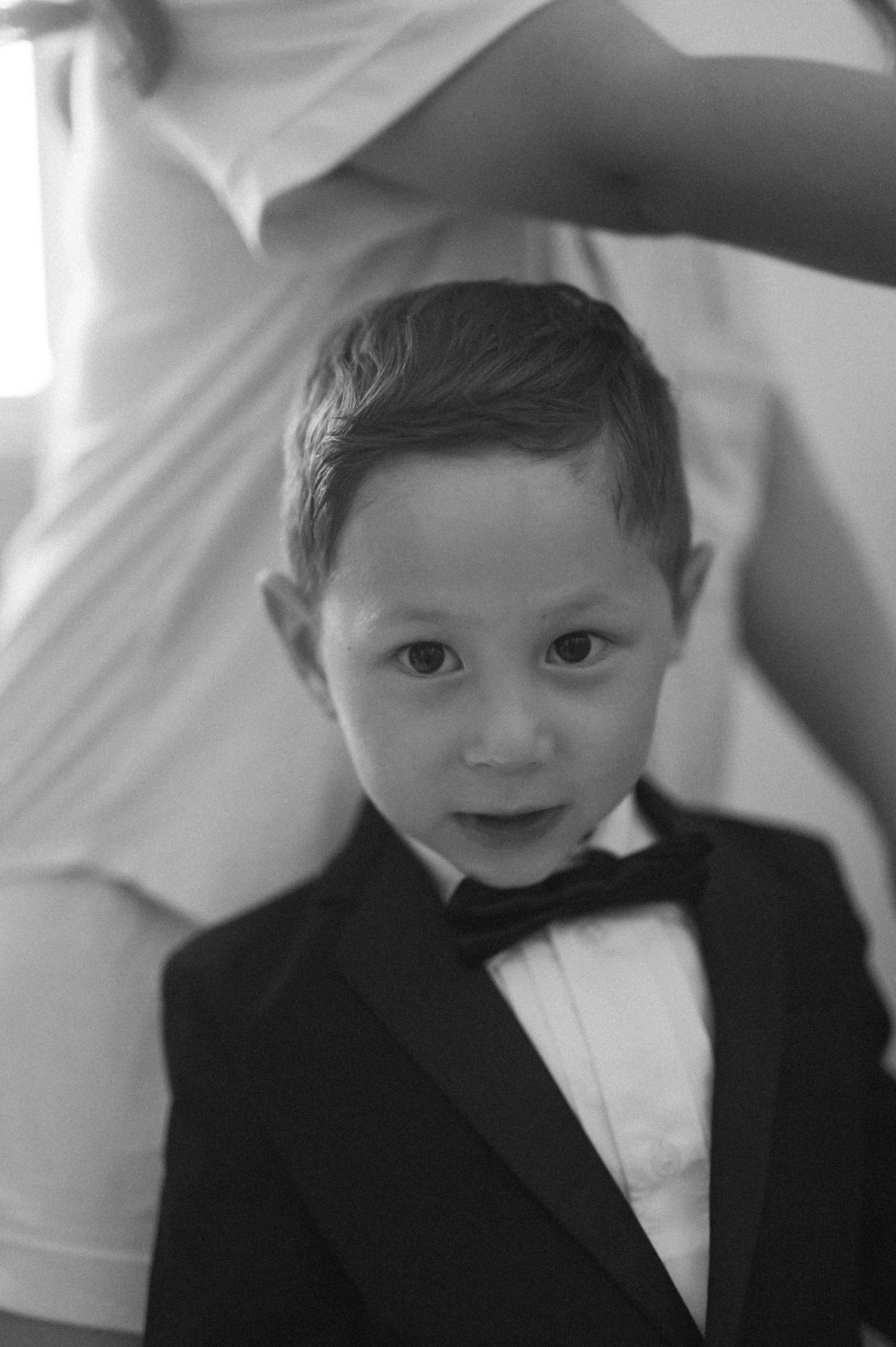 Adorable young boy in a black tuxedo with bow tie poses for a formal portrait in black and white.