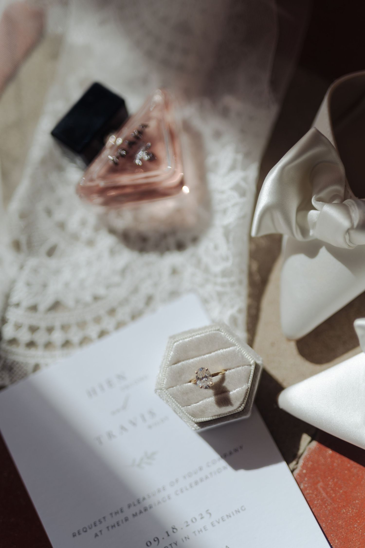 Wedding details including a perfume bottle, engagement ring box, invitation and white bridal shoes arranged on lace fabric.