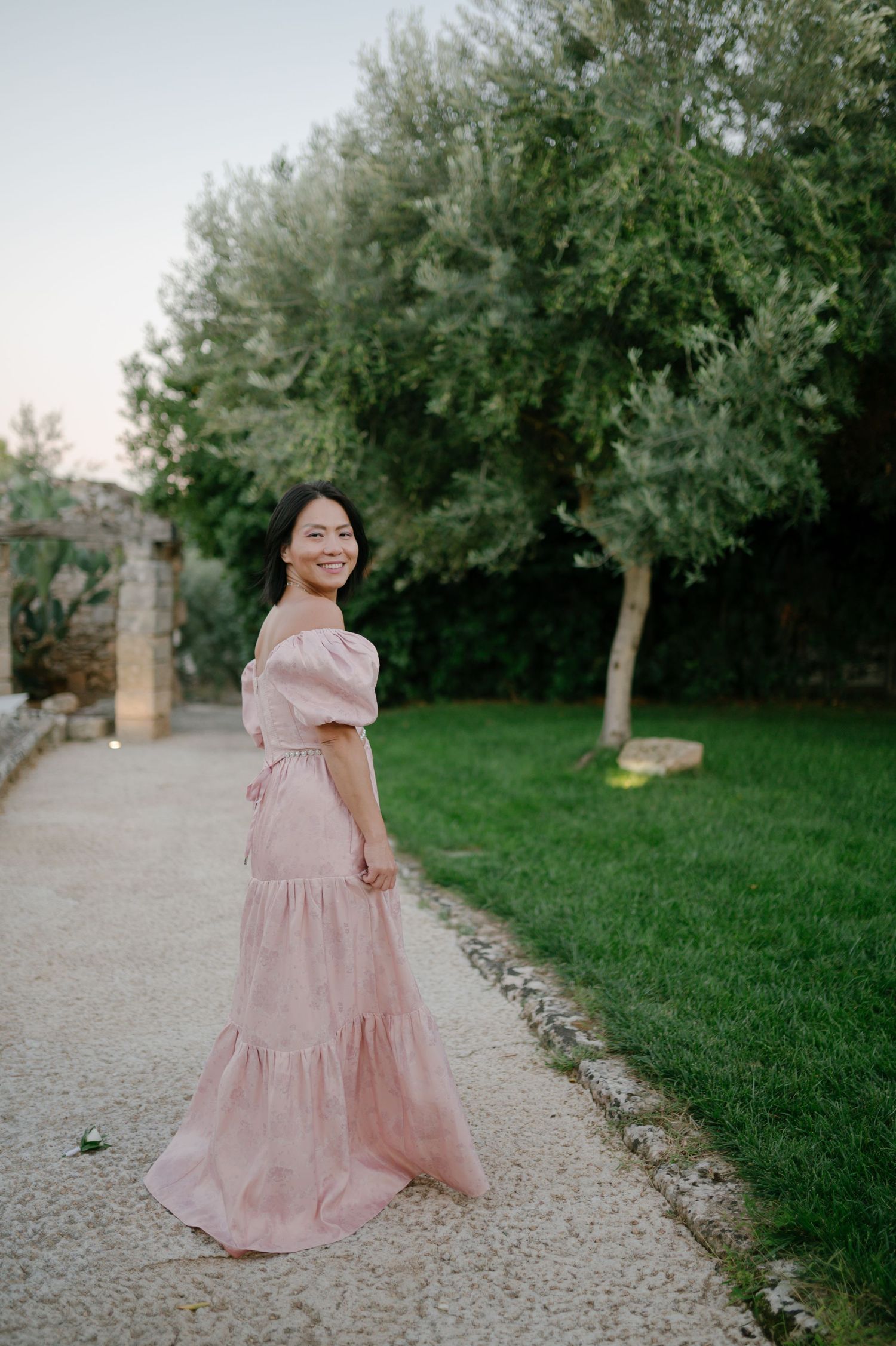 A person in a flowing pink dress walks along a gravel path surrounded by lush green trees and grass.