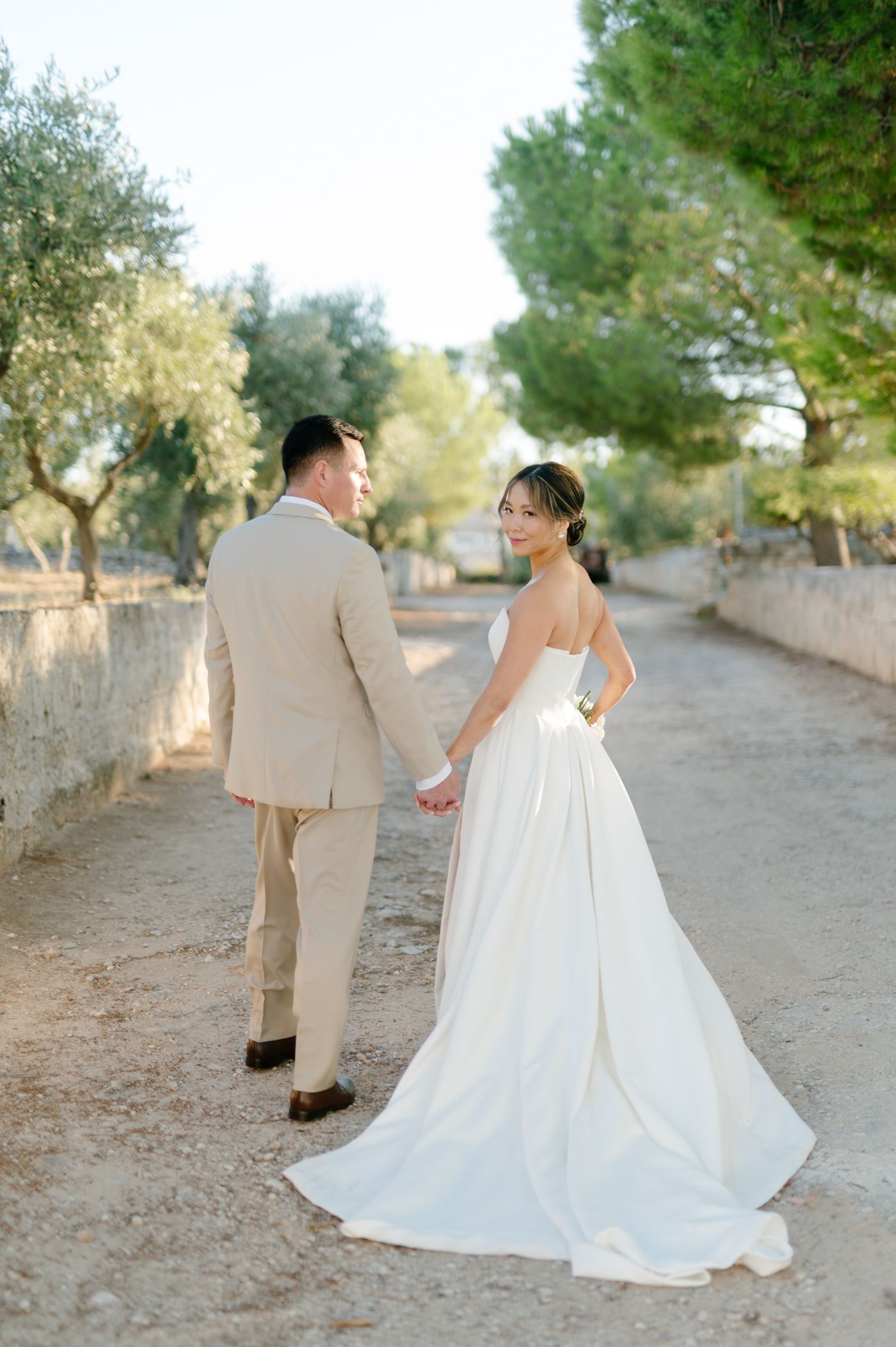A couple in wedding attire walking down a tree-lined path with stone walls on a sunny day.