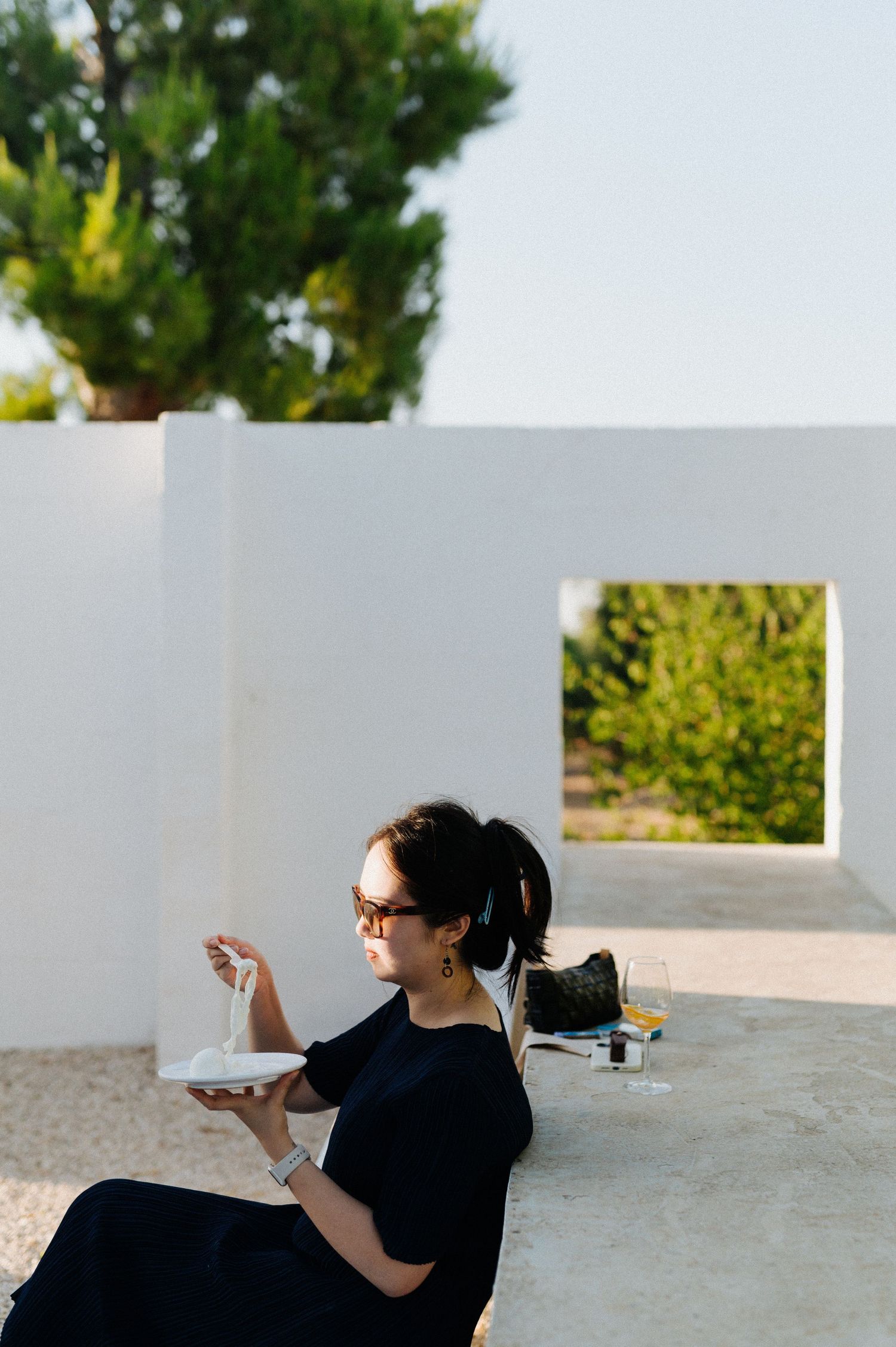Person in black shirt sits on white ledge outdoors with green foliage visible in background.