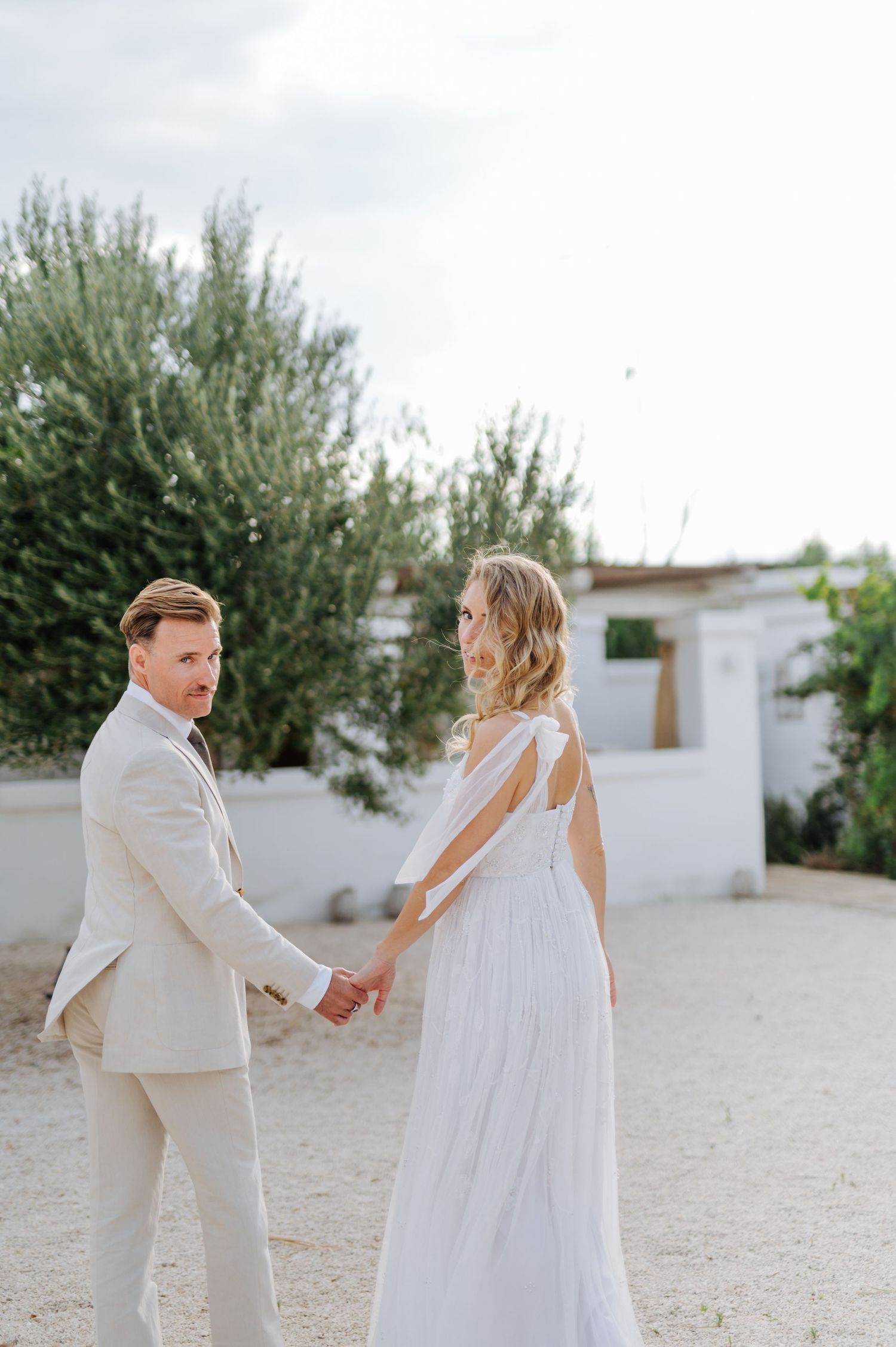 Couple in white attire dancing together on sandy ground near white buildings.
