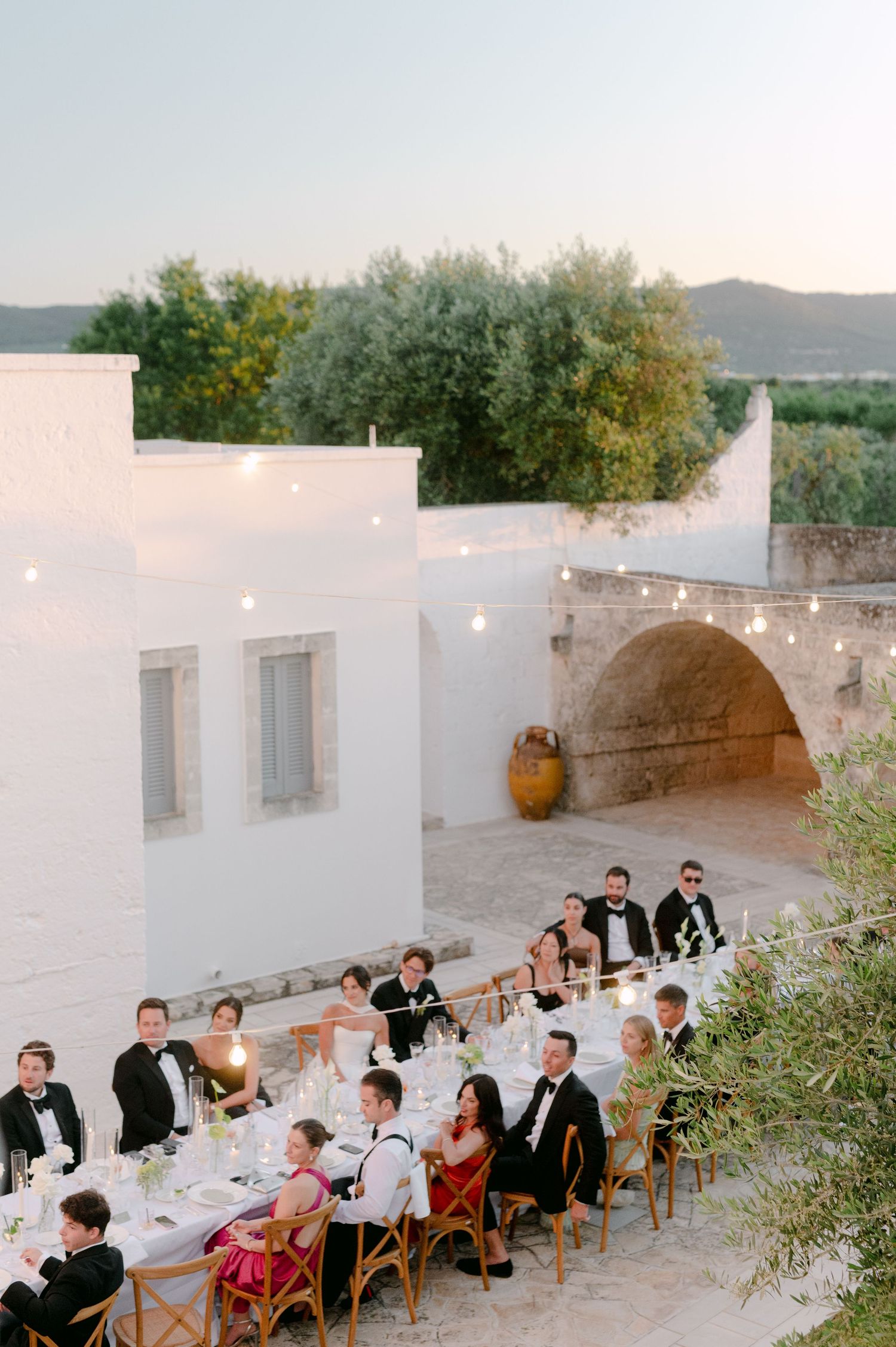 Outdoor wedding reception dinner at a whitewashed Mediterranean venue with string lights, long tables, and guests in formal attire.