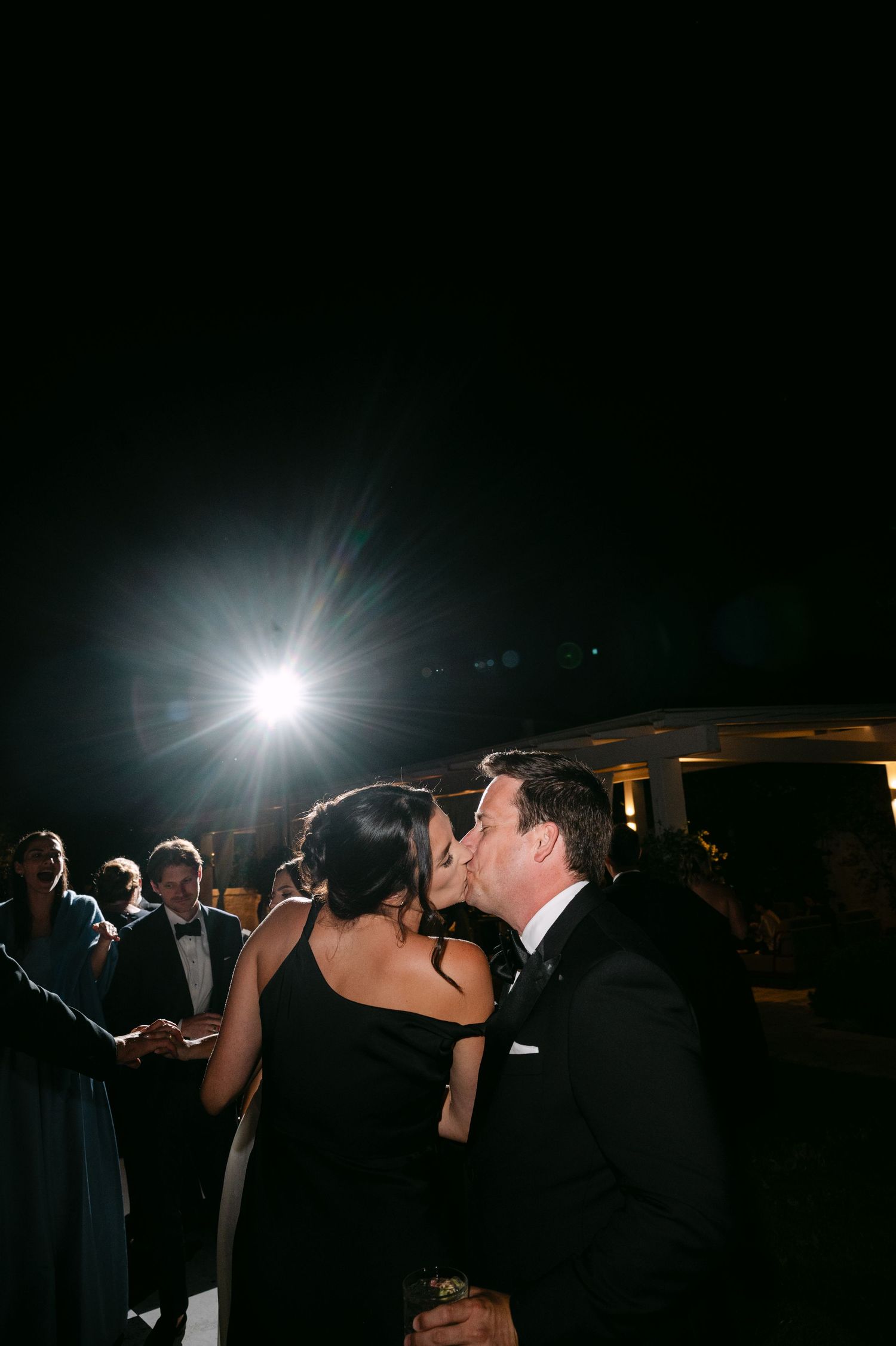 Couple sharing a romantic kiss at an outdoor evening wedding reception with guests and bright lights in the background.