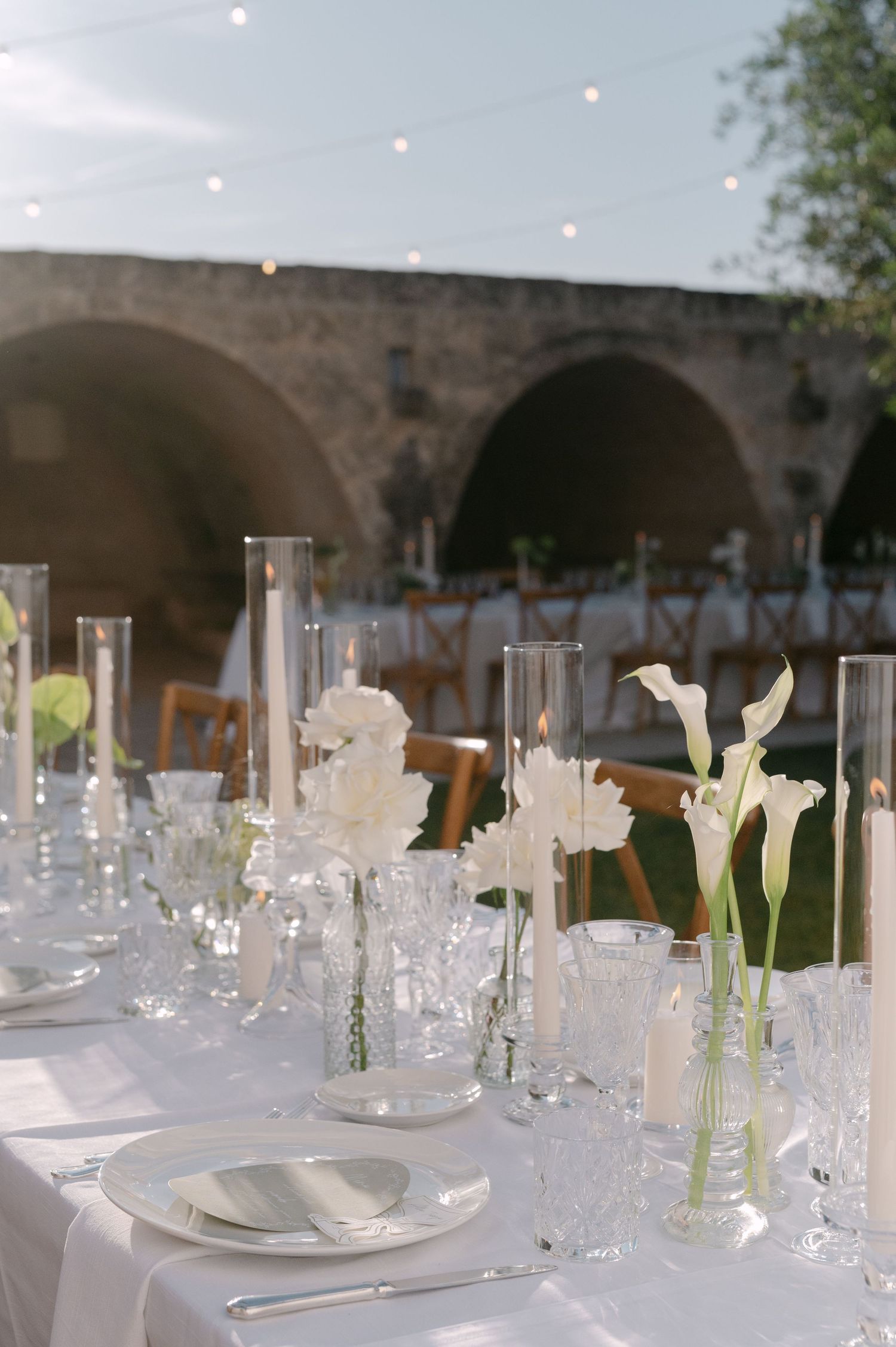 Elegant outdoor wedding reception table with white floral centerpieces, crystal glassware, and candles near a stone bridge.