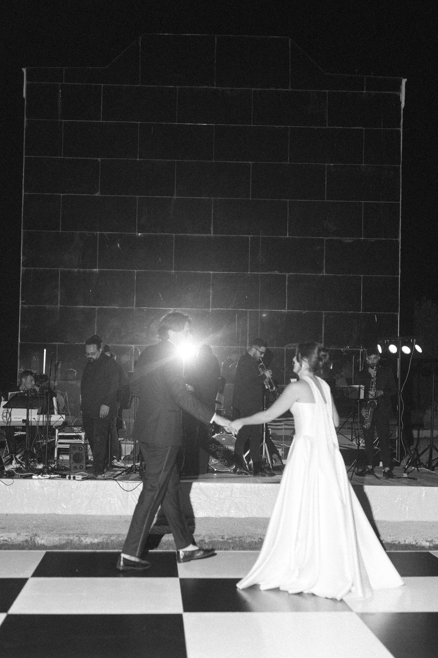 Black and white photo of a bride and groom sharing their first dance on a checkered floor with a live band performing behind them.