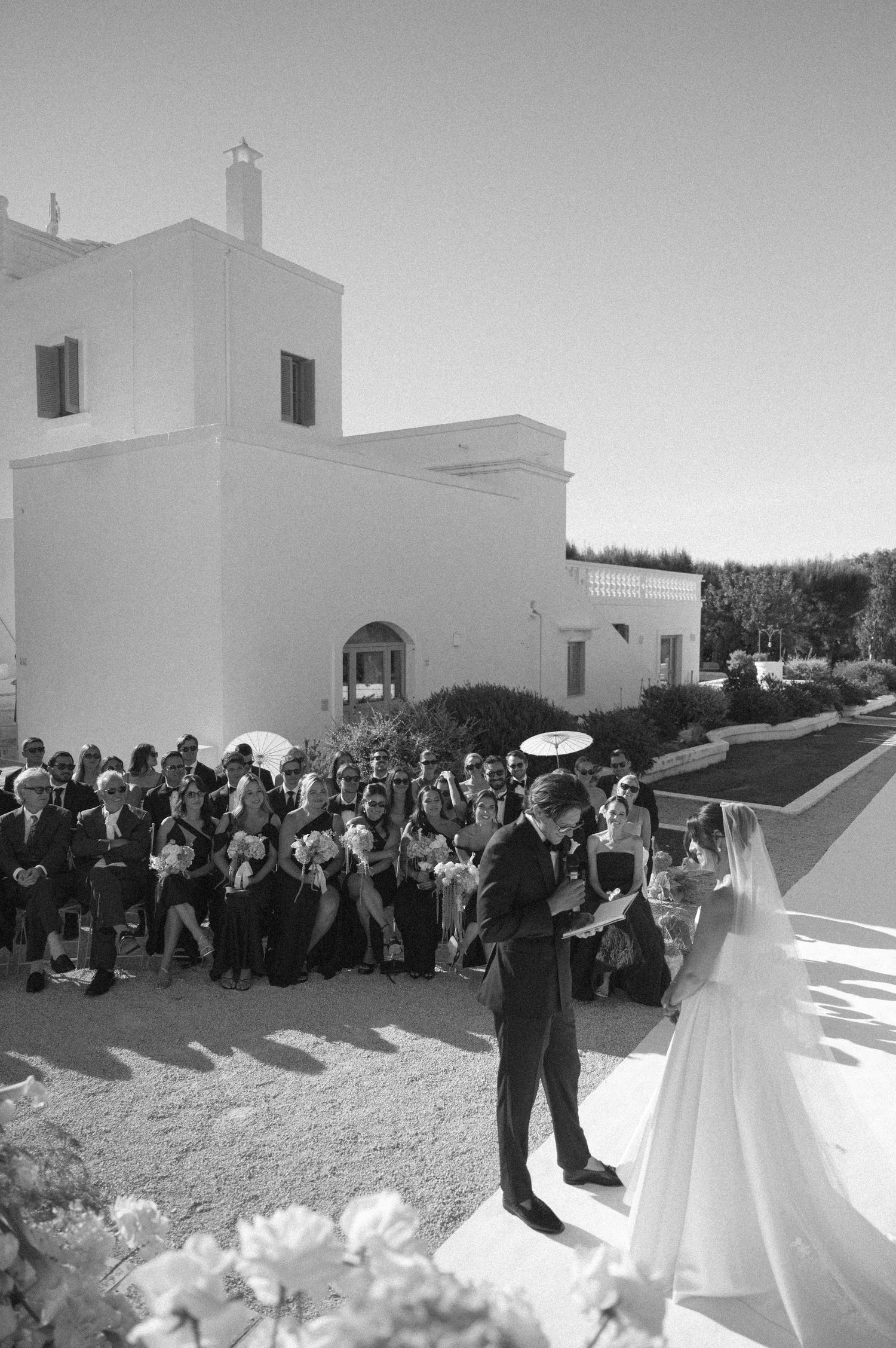 Black and white photo of a wedding ceremony outdoors at a white Mediterranean villa with guests seated.