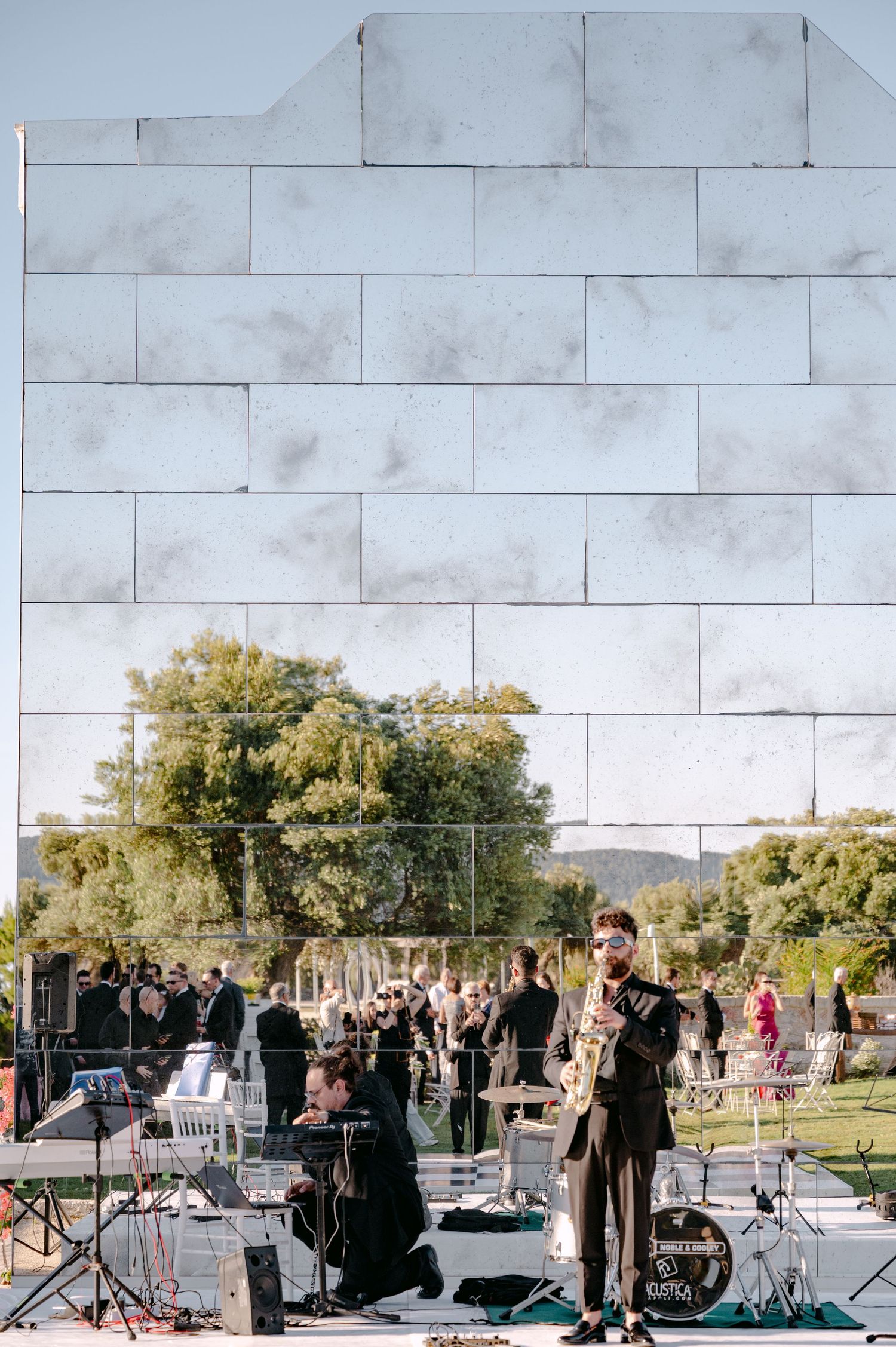 Jazz musician plays saxophone outdoors at an event, with a mirrored wall reflecting trees and guests behind.