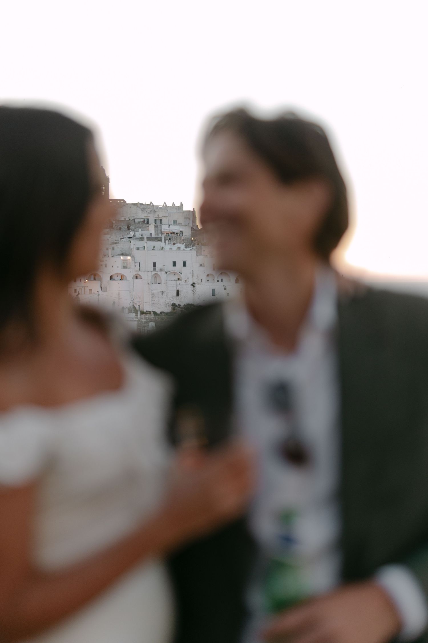 Blurred couple in wedding attire smiling with ancient hilltop town of Matera, Italy in soft focus background.