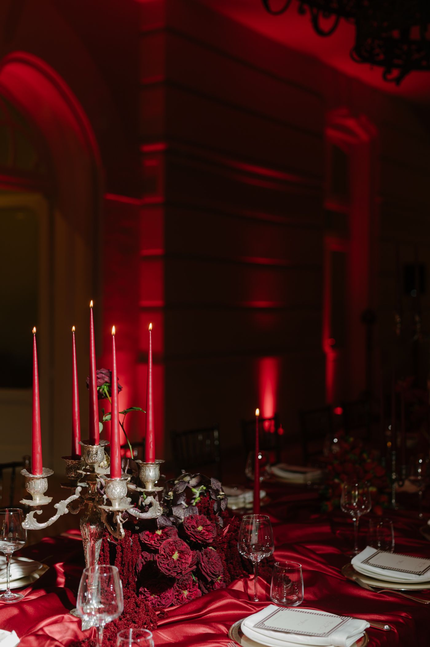 Elegant table settings with red tablecloths and tall white candles create a dramatic atmosphere under red-tinted lighting.