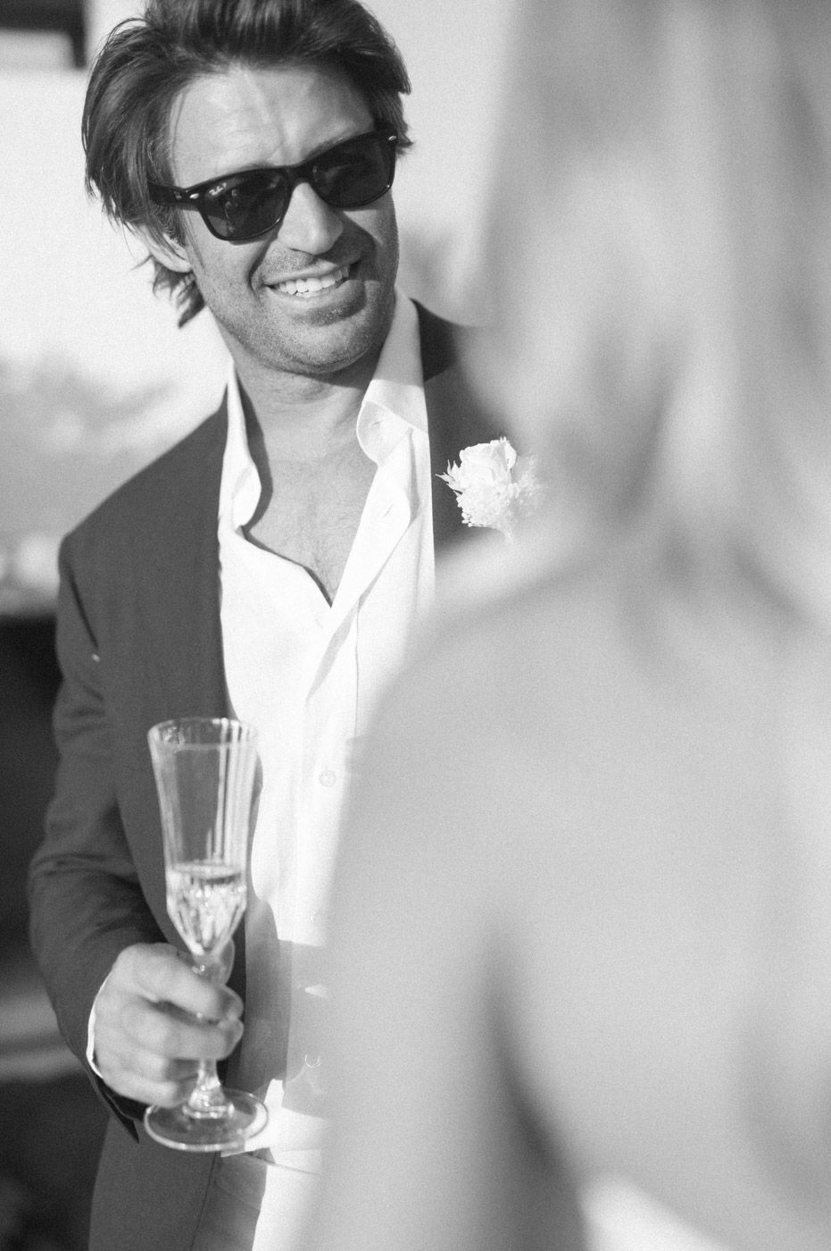A person in sunglasses and a black suit smiles while holding a champagne glass at a formal event.