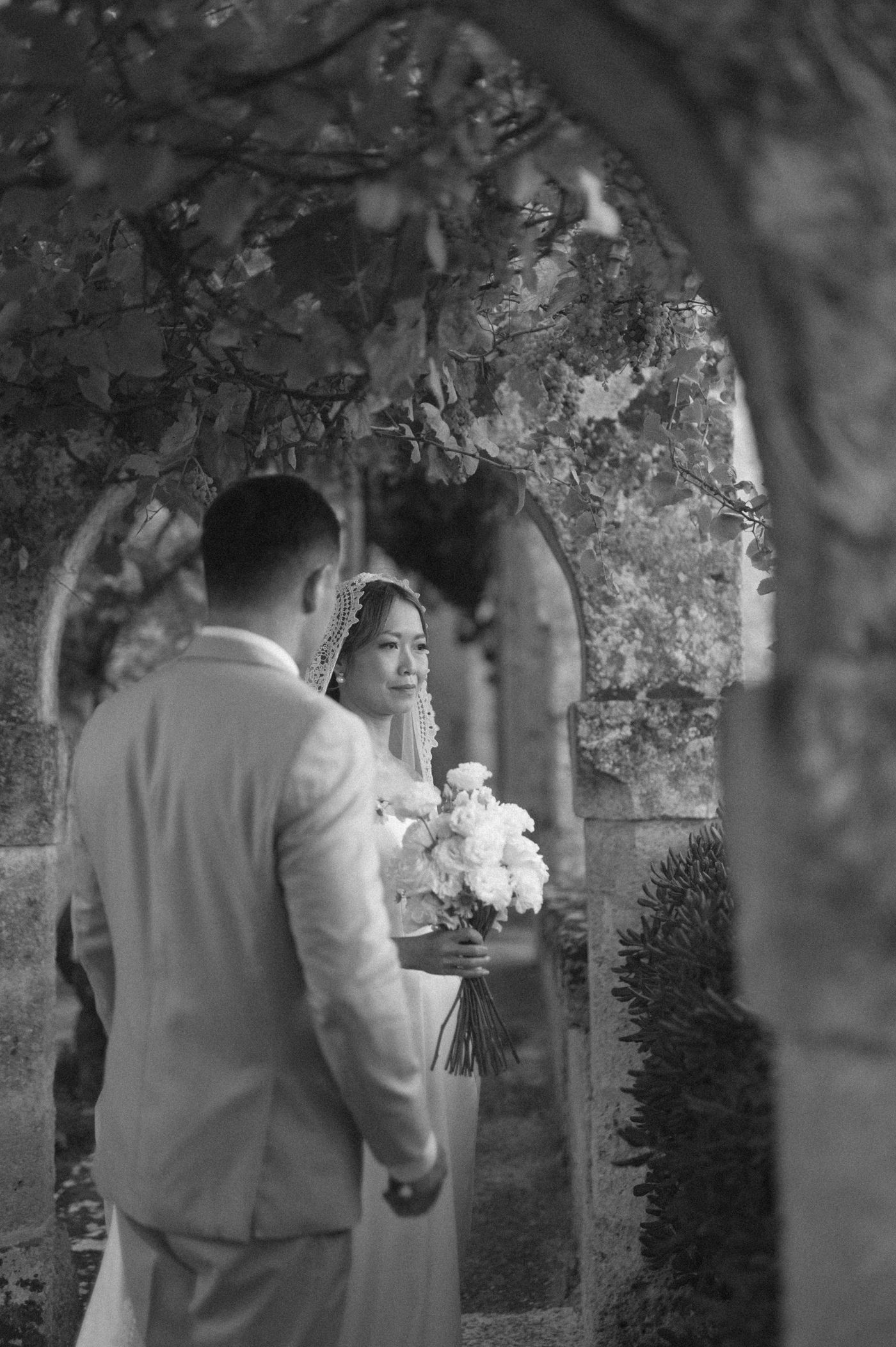 A romantic black and white wedding photograph showing a couple in an elegant garden archway with flowers.