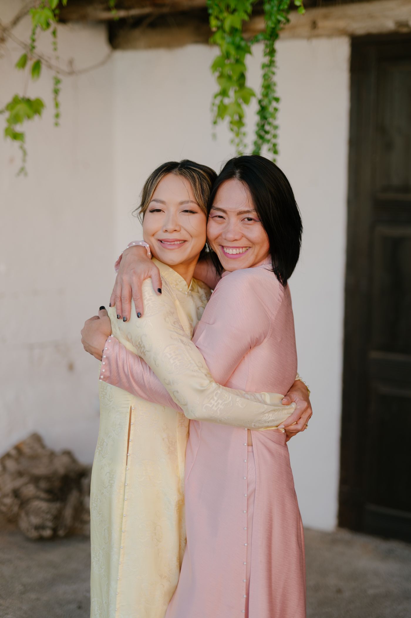 Two people wearing yellow and pink dresses embrace warmly while smiling in front of a white wall with green foliage.