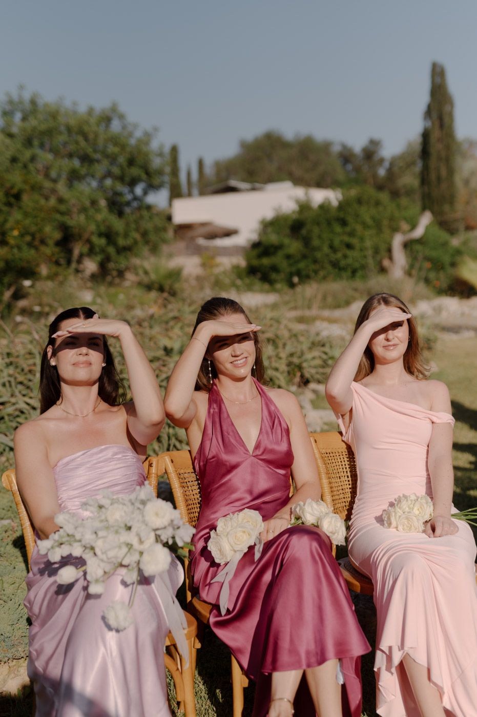 Bridesmaids in pink dresses shielding eyes from sun while holding white bouquets.