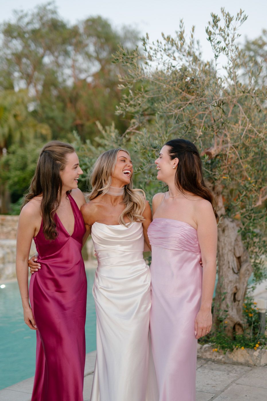Three women in formal dresses laugh together during a wedding photoshoot by a lake.