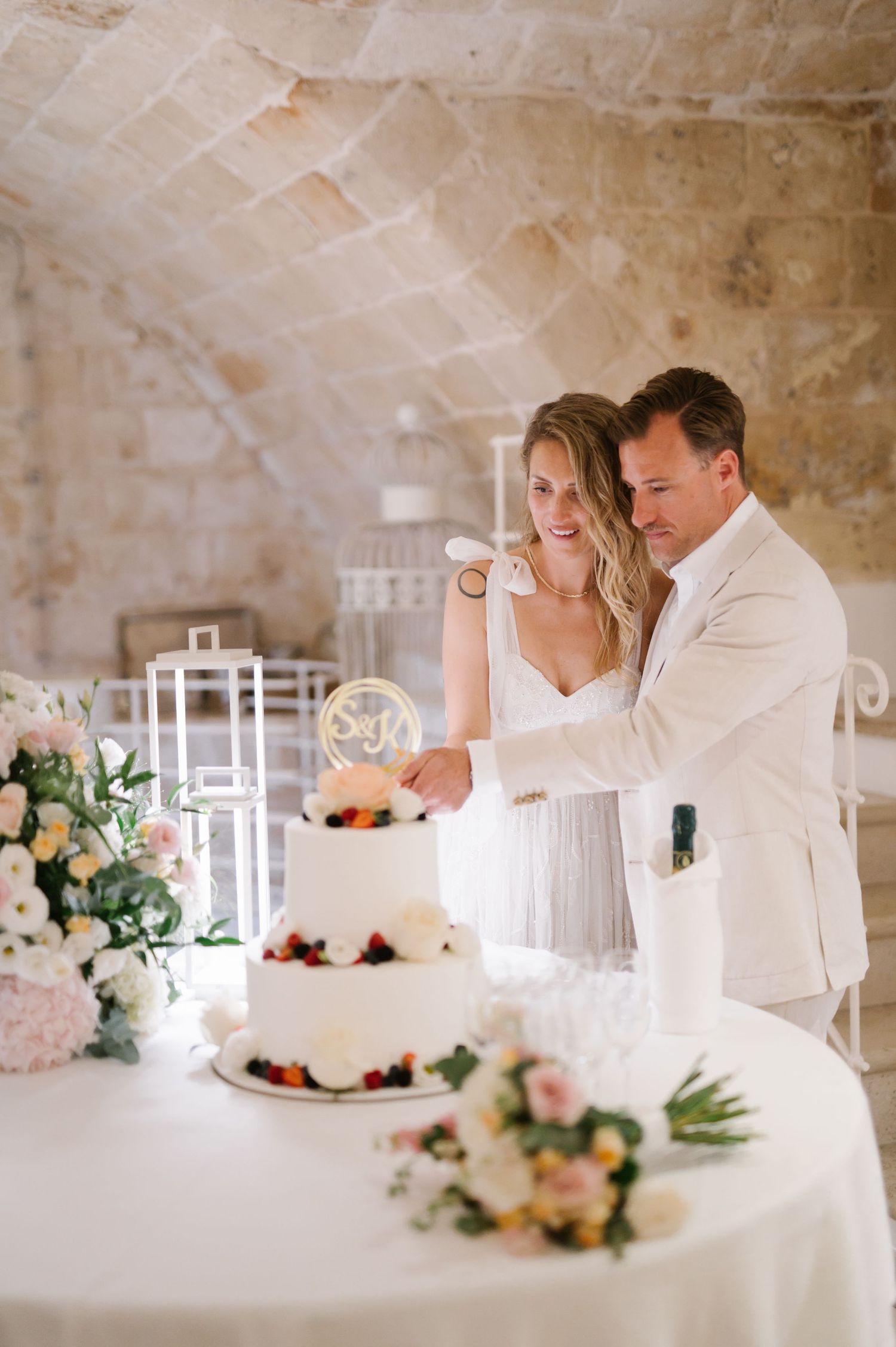 Couple cutting their white wedding cake decorated with fresh flowers at a rustic stone venue.