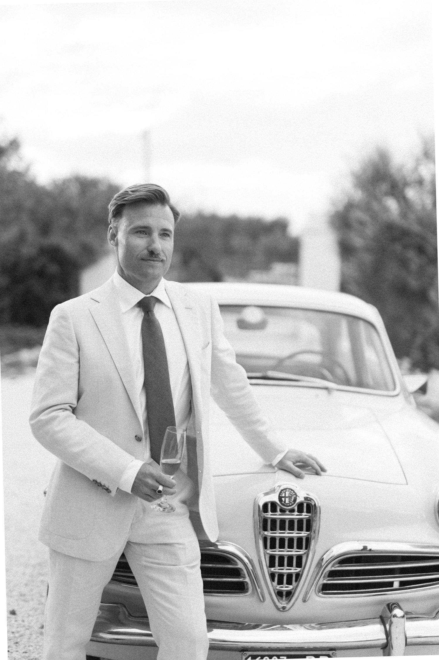 Black and white portrait with classic Alfa Romeo car and elegant attire.