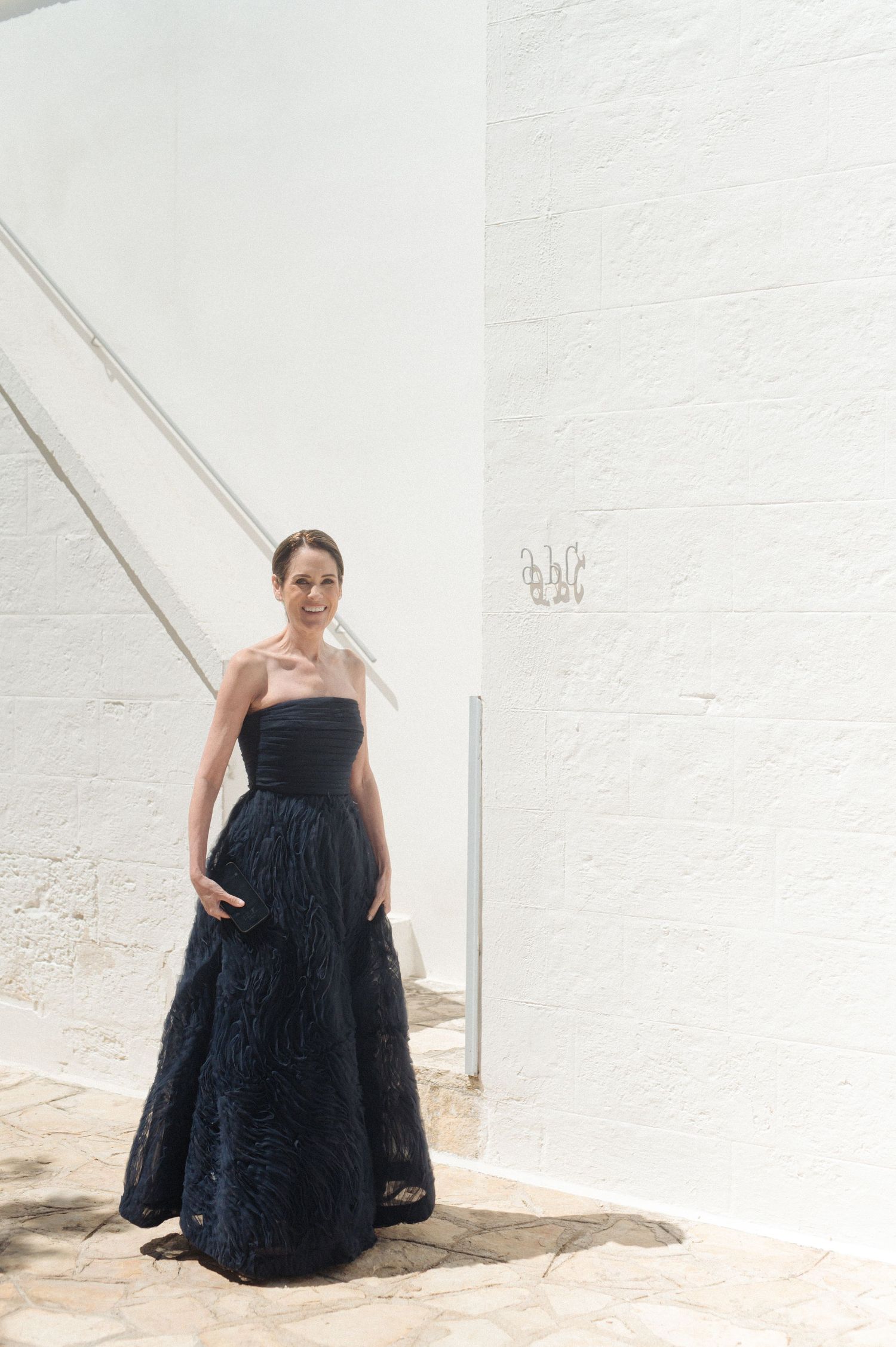 Woman in elegant navy blue strapless ball gown poses against white textured wall with staircase backdrop.