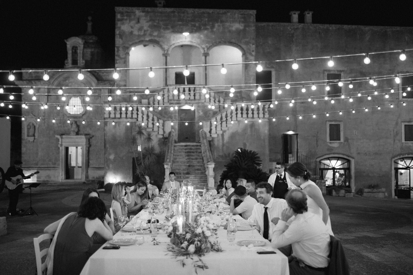 Guests dine at an elegant outdoor table under string lights in front of a historic stone building at night.