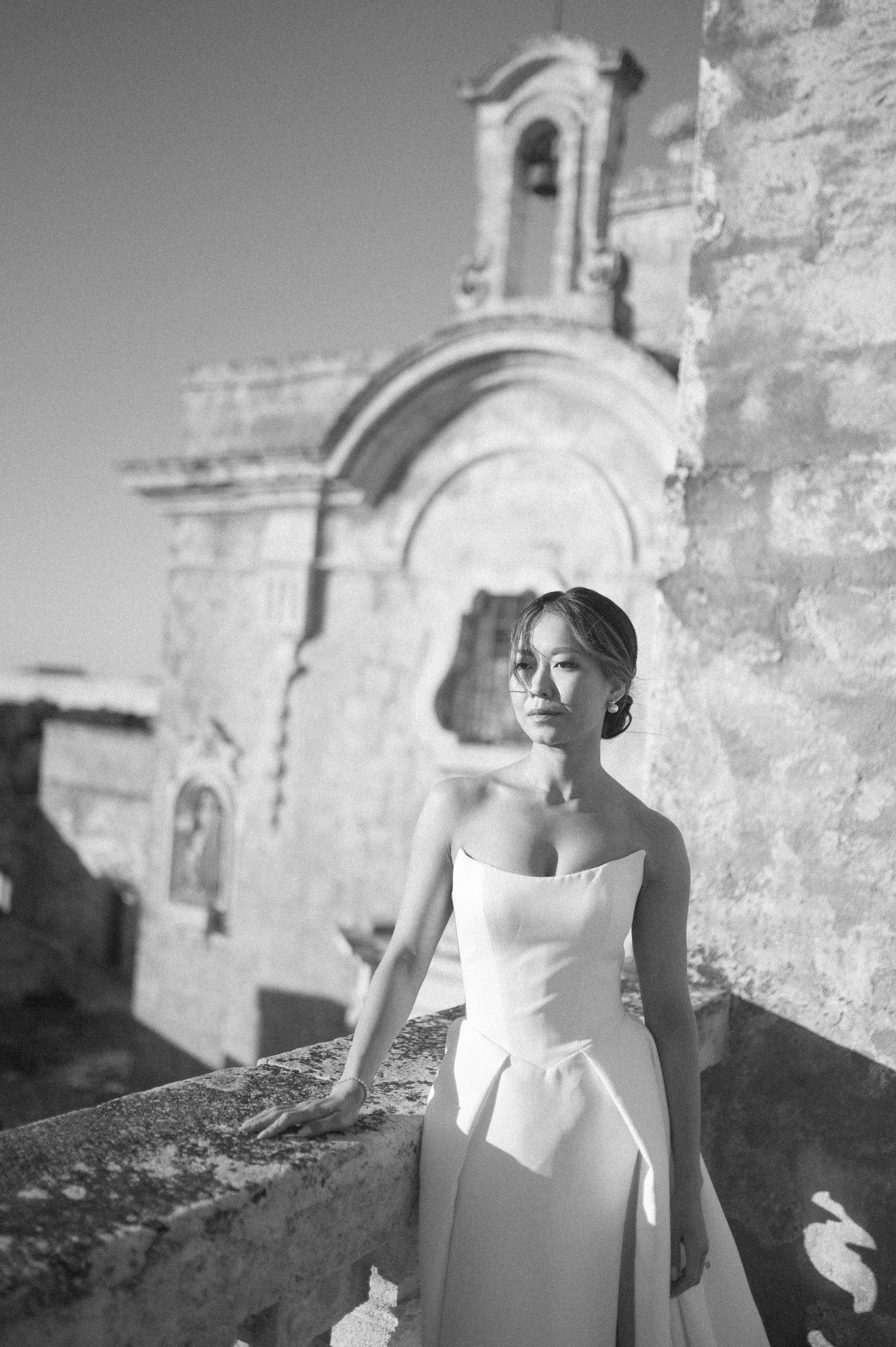 A black and white photo series showing a person in a white dress posing against stone architecture in bright sunlight.