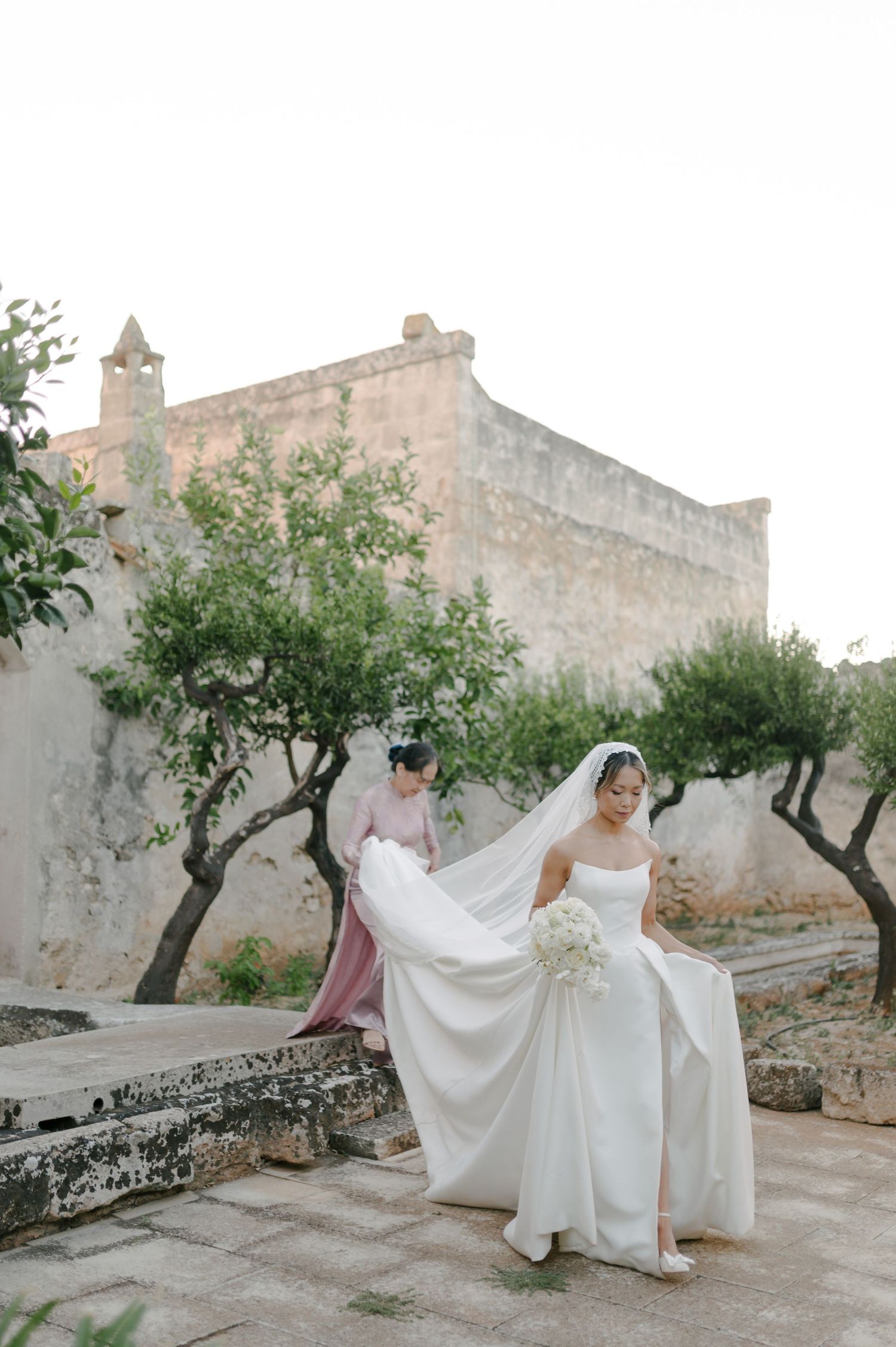 A flowing white wedding dress is elegantly displayed against stone walls and olive trees in a Mediterranean setting.