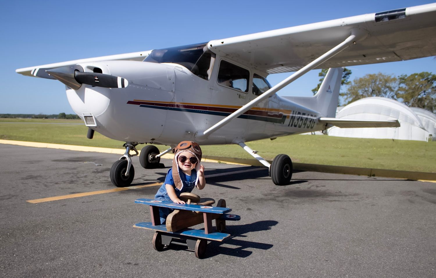 Young child sitting on a toy airplane on a tarmac with a real Cessna aircraft parked behind them.