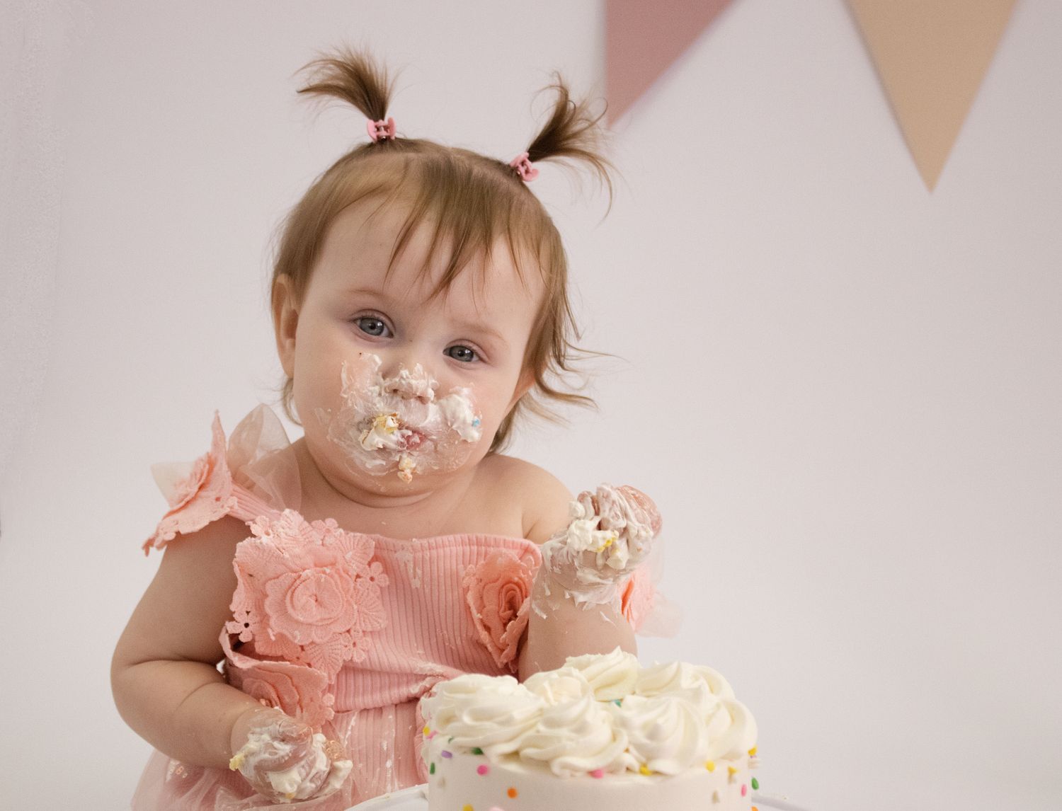 Baby girl in pink dress smashes first birthday cake, face covered in frosting, with festive bunting in background.