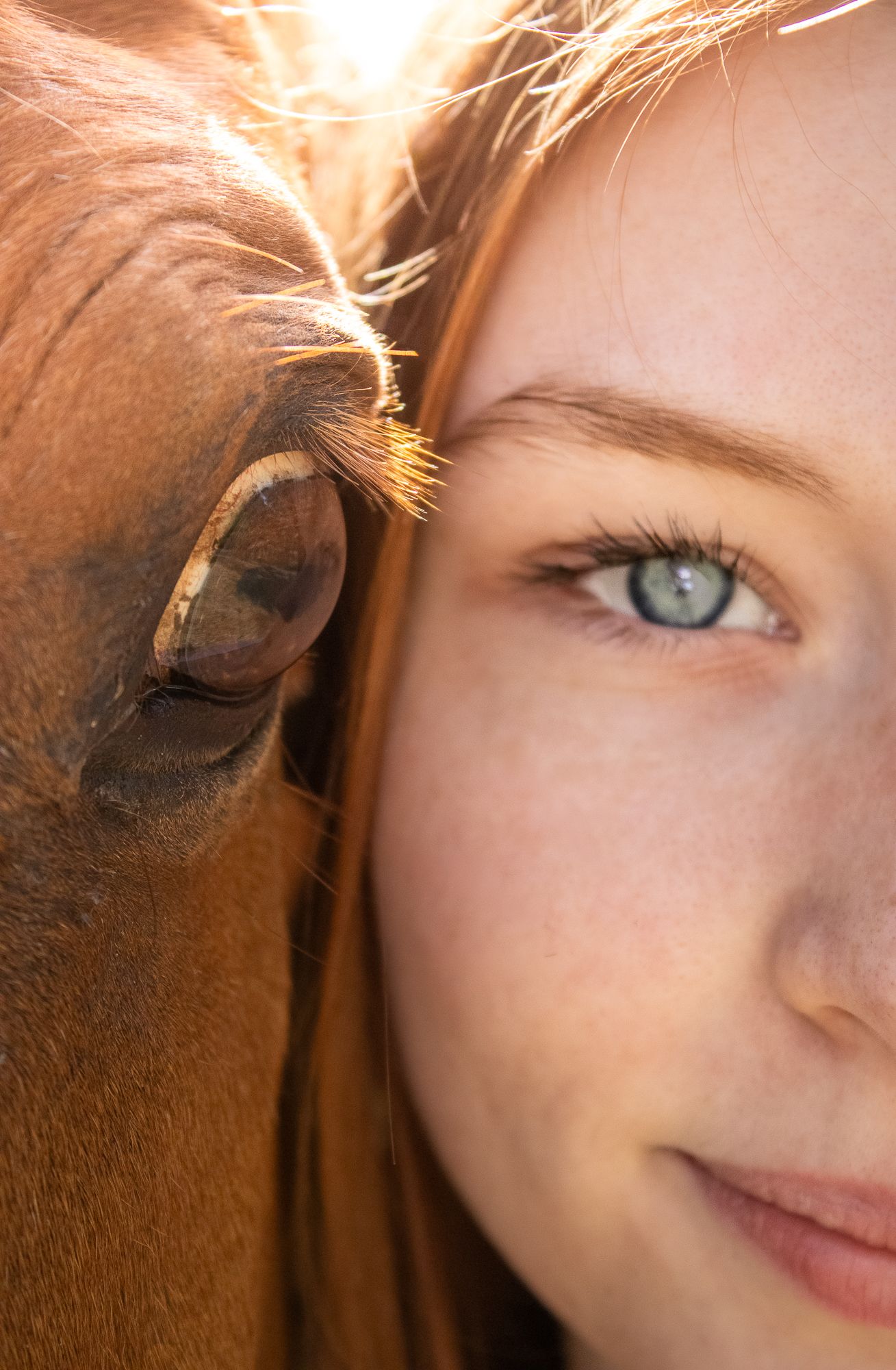 Close-up portrait of a young redhead girl with green eyes beside a brown horse, both facing the camera.
