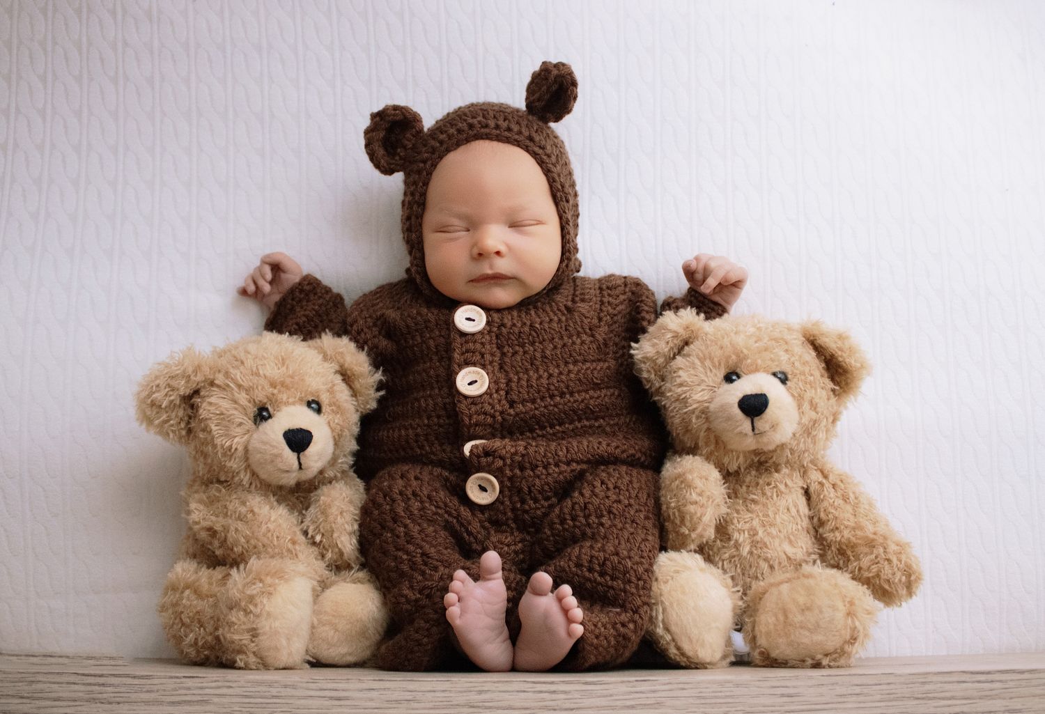 Sleeping newborn baby in brown knit bear outfit sits between two teddy bears for an adorable photo.
