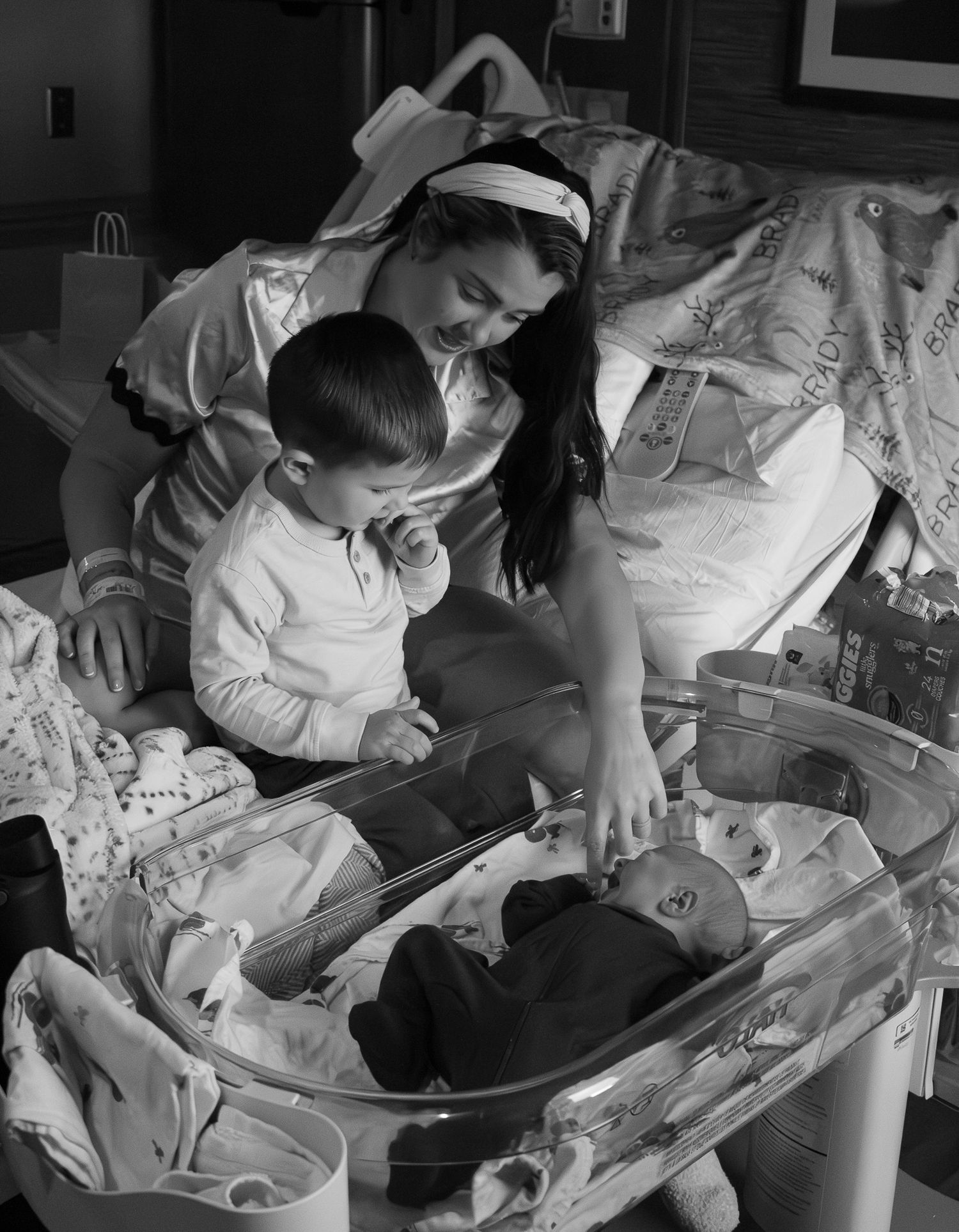 Black and white photo of a mother in hospital bed with toddler son looking at newborn baby in bassinet.