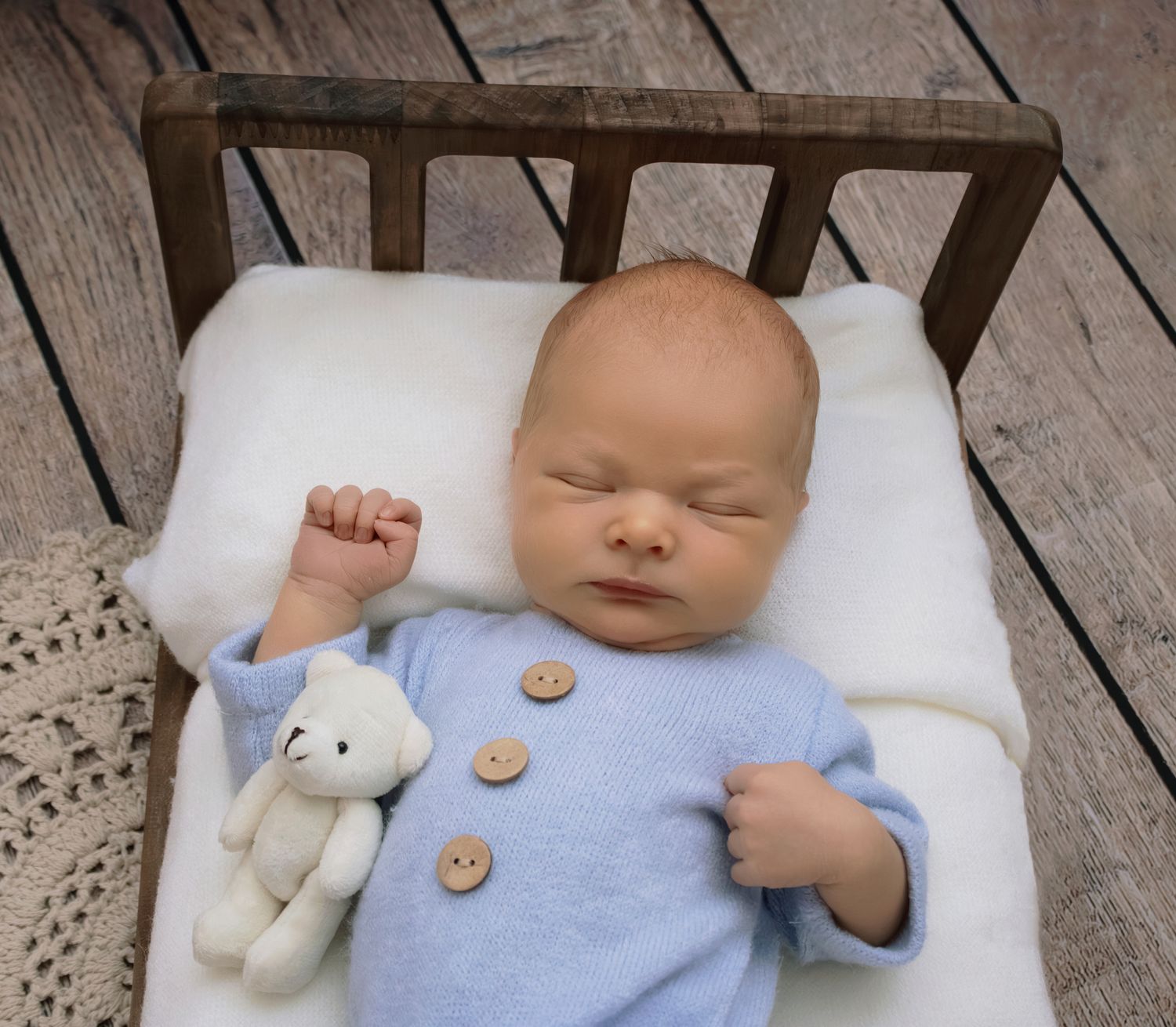 Sleeping newborn baby in blue knit onesie lying in small wooden bed with white teddy bear.