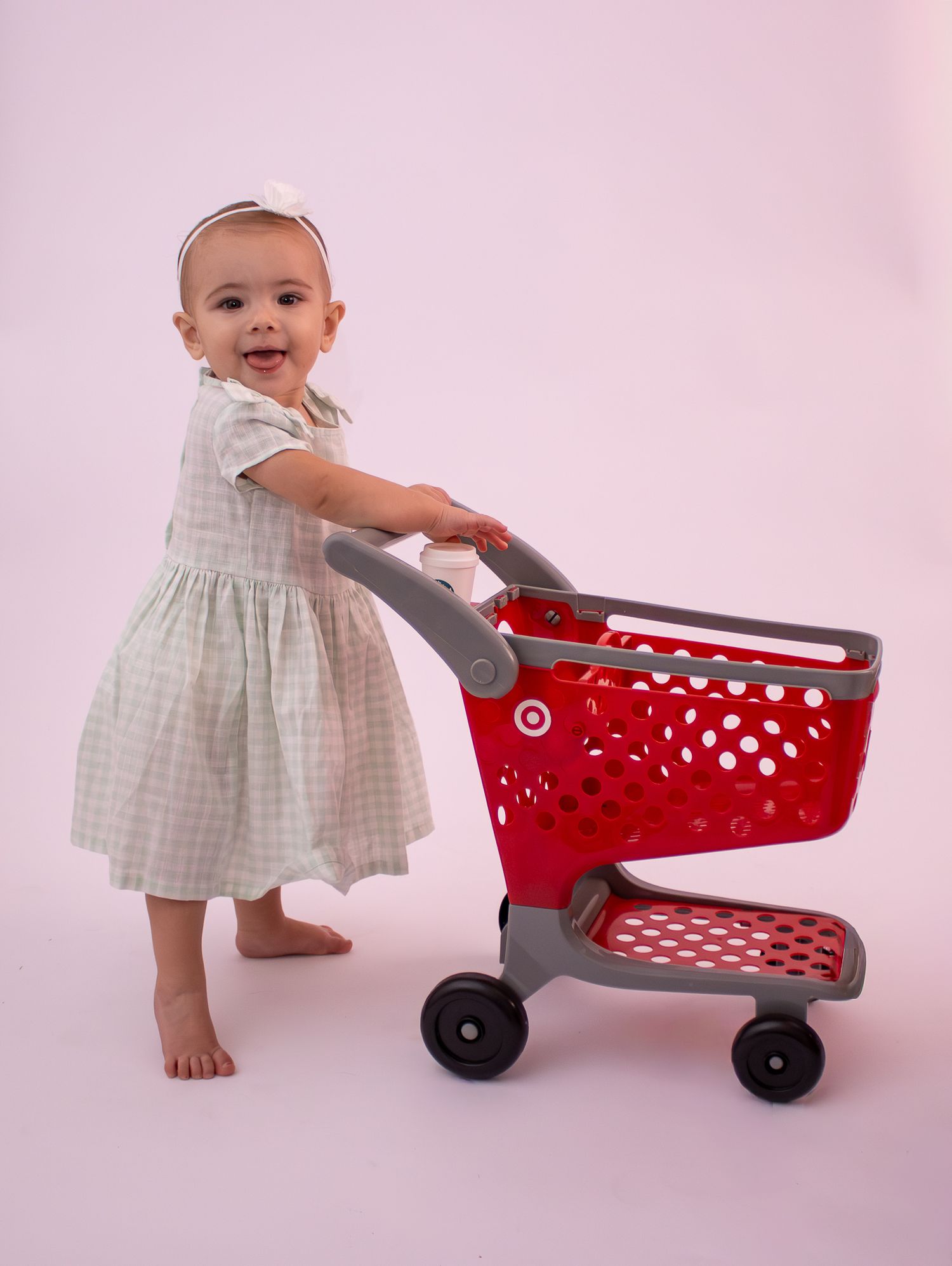 Baby girl in gingham dress pushes red Target toy shopping cart, smiling at camera on pink background.